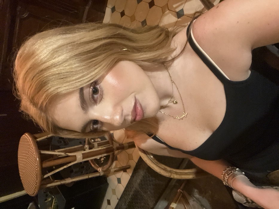 A blonde woman wearing a black tank top poses. A gold necklace with a pendant is visible on her chest. She has fair skin and a slight smile. Her hair is styled in loose waves. On her left wrist, she wears several gold bracelets. In the background, a stool with a round woven seat is visible, along with tiled flooring in shades of beige, brown, and blue. The lighting suggests an indoor setting with artificial light.