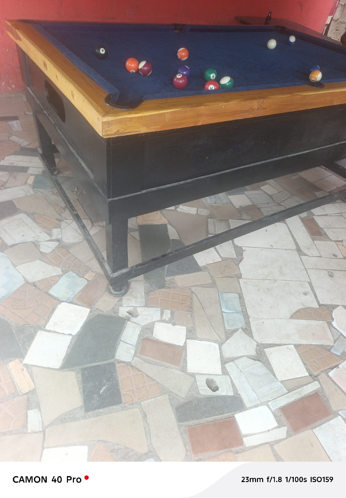 The image displays a pool table positioned in an indoor or covered outdoor setting. The table features a dark blue felt surface, a light brown wooden frame, and a black body supported by black metal legs. Numerous numbered pool balls, including solids (red 3, orange 5, green 9, blue 7, yellow 1), striped balls (red and white, orange and white, green and white 6, blue and white), and the black 8 ball, along with a white cue ball, are scattered across the playing area.
Behind the pool table is a prominent red wall. Faintly visible on a surface against the wall in the upper right corner is what appears to be a dark bottle.
The floor beneath and in front of the table is a distinctive mosaic made of irregularly shaped, multi-colored ceramic tiles. These tiles are in various shades of white, light blue, brown, grey, black, and reddish-orange, appearing somewhat worn.
No people are present, but the arrangement of the balls suggests a game was recently played or is in progress. The lighting indicates it is daytime. The rustic appearance of the table and the unique tiled floor hint at a casual establishment or community space, consistent with the location context of Jos, Nigeria.
Visible text at the bottom left reads "CAMON 40 Pro," indicating the camera model. At the bottom right, the camera settings "23mm f/1.8 1/100s ISO159" are displayed.
