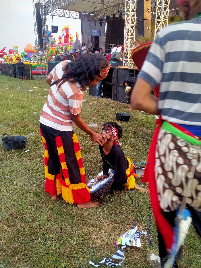 The image captures an outdoor scene at an event in Glonggong, Indonesia, taking place on a grassy field. In the immediate foreground, three individuals are present. A person with long, dark hair, wearing a white and brown striped shirt and black pants adorned with prominent red and yellow fringed sections, stands barefoot. This individual is actively manipulating the open mouth of a child who is seated or kneeling before them. The child wears a black long-sleeve shirt and yellow pants; their face is painted with stylized black and white makeup around the eyes and mouth, and a red and white patterned bandana is on their head. The child holds a patterned prop, consistent with a hobby horse head (Kuda Lumping prop), featuring white, blue, and black elements with dark, feathery attachments. To the far right, partially visible and out of focus, another individual is seen from behind, wearing a blue and white striped shirt, colorful sashes around the waist, and a red clown nose. In the background, a large stage structure constructed from metal trussing is prominent, flanked by stacked black sound speakers. Individuals are discernible on and near the stage. To the far left of the stage, several large, colorful inflatable amusement rides, including a red creature-shaped one, are visible. The ground consists of green grass with scattered pieces of debris and litter. A black plastic bucket rests on the ground to the left of the central figures.