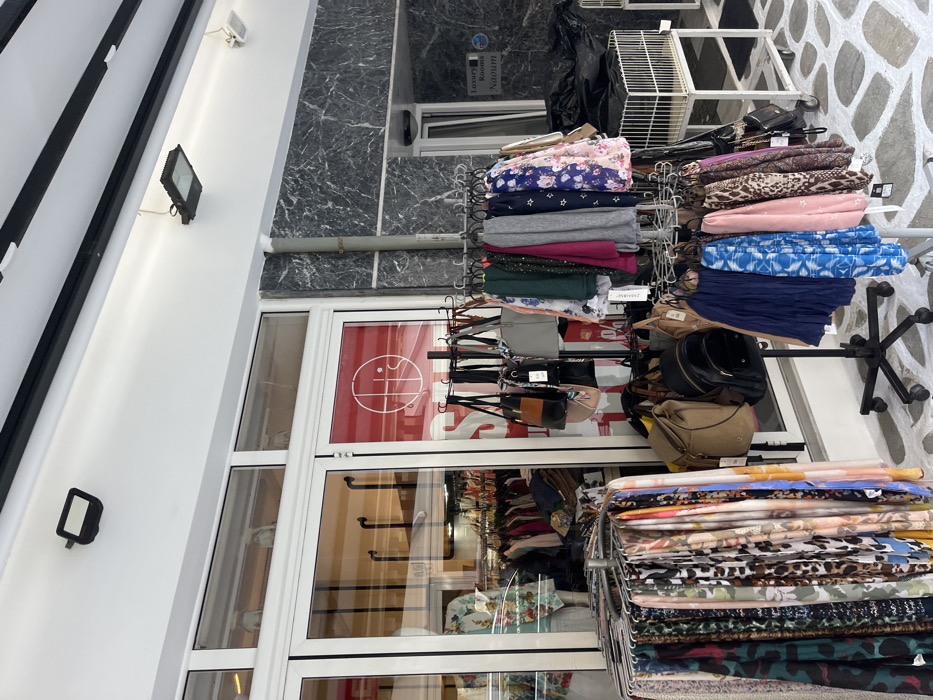 The image shows a storefront with a rack of scarves in front of it. The storefront has a white facade with a black marble trim. The storefront window displays a sign for a sale. The storefront window also has a display of purses and handbags. There are two lights mounted on the exterior of the storefront, one above the window and one on the exterior wall above the doorway. The storefront is located on a cobblestone street. There are no people present in the image. The location is Limenaria, Greece, at coordinates (latitude: 40.626, longitude: 24.575).