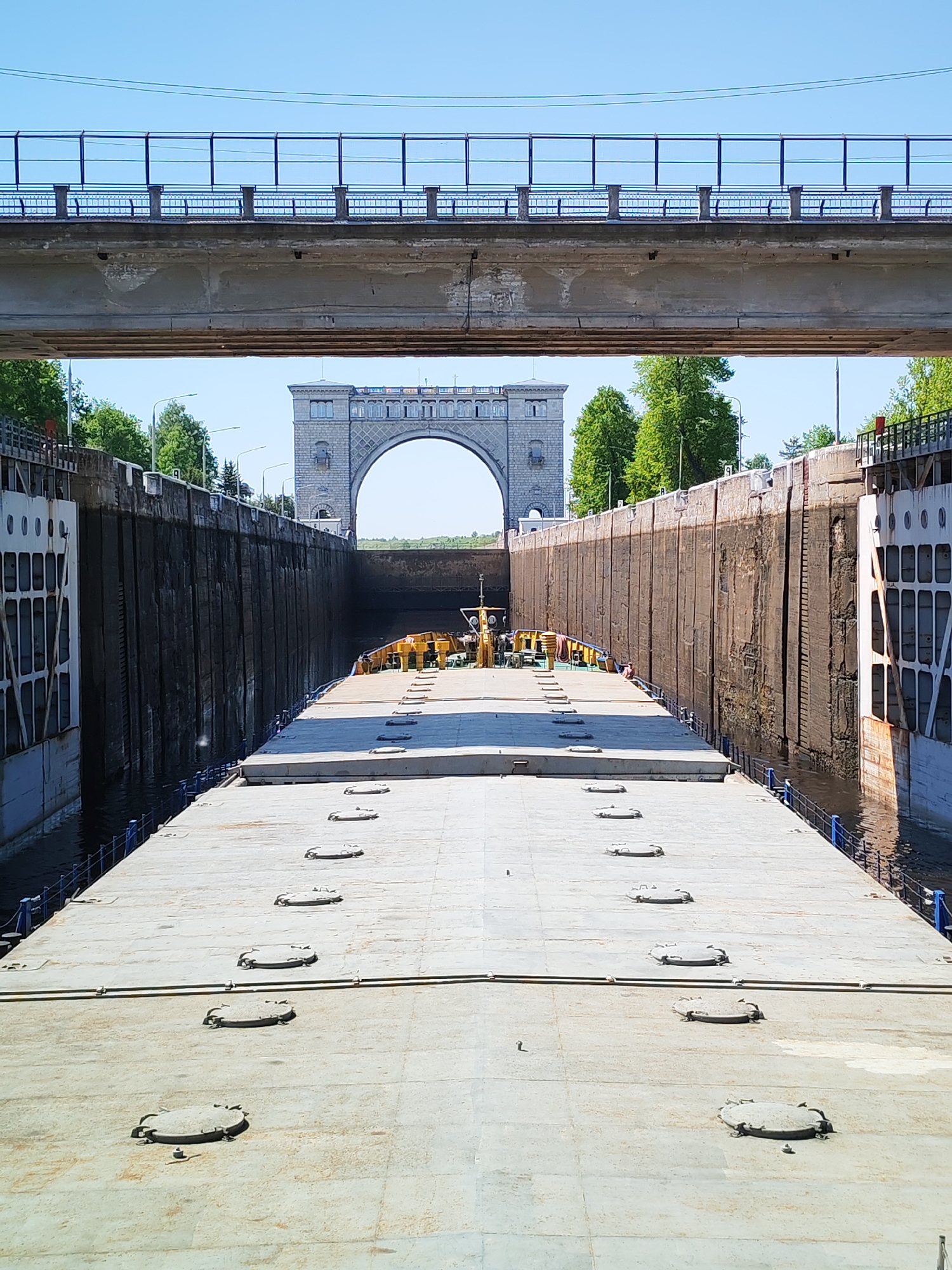 A barge is in a river lock in an unidentified city. The lock walls are made of concrete and steel. The barge is long and flat with many circular hatches on its deck. There is a bridge over the lock at the foreground and a large archway and tower is visible at the far end of the lock. Trees line the sides of the lock. The weather is clear and sunny.