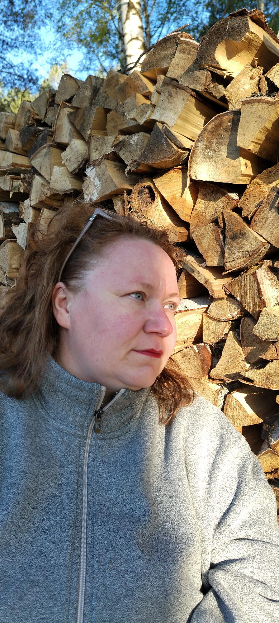 A woman with light skin and reddish hair, wearing a gray zip-up sweatshirt and glasses perched on her head, is positioned in front of a large stack of firewood. The firewood is composed of split logs, their surfaces displaying wood grain and textures. The environment appears to be outdoors, with hints of foliage and a tree trunk visible in the background. The location is Зипуново, Russia.