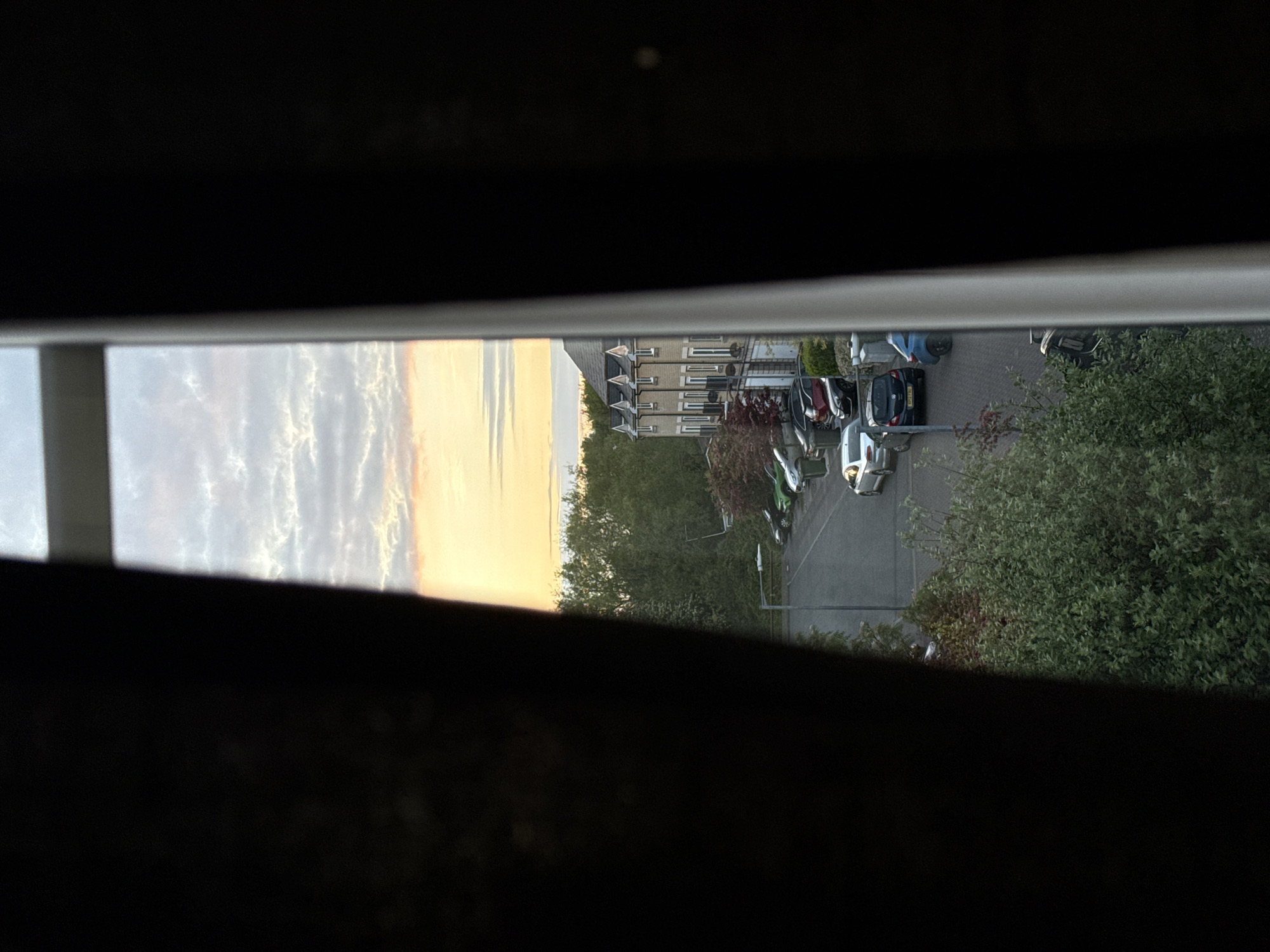 The image shows a view through a partially open window. The scene outside includes a sky with clouds illuminated by a setting sun, a row of houses, trees, a street with parked cars, and greenery. The location cannot be determined based on this image. There are no visible people or actions taking place.
