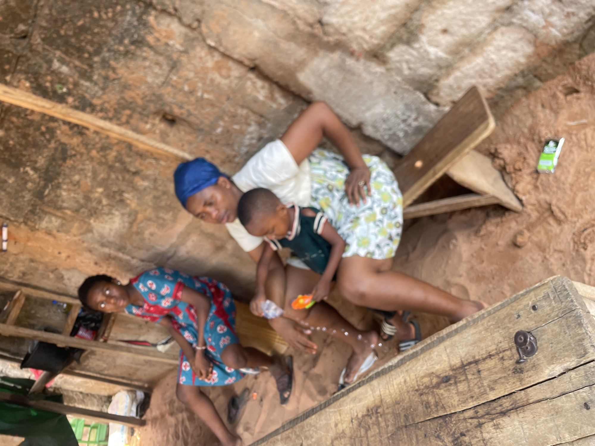 The image shows an outdoor scene in Nkpor, Nigeria. An adult woman in a white shirt, floral skirt, and blue head covering is holding a young child who wears a dark top with white trim and is playing with a toy gun. Another young person in a blue and red patterned dress sits nearby. The adults and children are on or near a rough wooden platform. The background consists of earth, rock, and crude wooden structures.