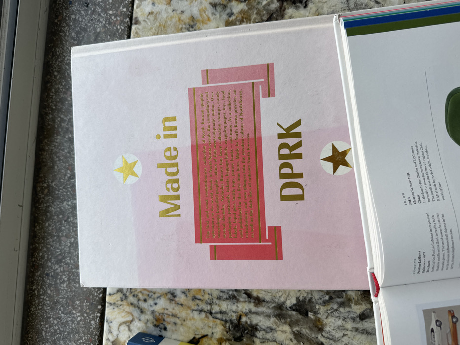 The image displays an overhead view of two hardcover books resting on a speckled light brown and grey granite or quartz countertop.
The primary subject is a closed book occupying the upper and central portion of the frame. Its cover is predominantly light pink, fading towards white at the edges, and features gold-colored text. The title "Made in DPRK" is prominently displayed. A rectangular pink block with gold outlining in the center of the cover contains descriptive text regarding the book's content, detailing a collection of North Korean graphic art, including items like stamps, candy wrappers, postcards, and labels. Two gold five-pointed stars, each set within a white circular background, are present on the cover: one centered near the top and another near the bottom.
Below this, a second hardcover book is open, with its pages partially visible at the bottom of the frame. The right-hand page displays printed text that includes "Charles Eames - 1958" and describes an "Eames Fibreglass Chair" with a "rocking base." A small portion of a red ribbon bookmark protrudes from the spine area of this open book.
To the far left, a small object with blue and yellow colors is partially visible on the countertop. The background above the countertop includes a dark horizontal strip, possibly a window sill or ledge, with a blurry white area above it.
No individuals are present in the image, and no actions are depicted other than the static arrangement of the objects. The location of Tel-Aviv, Israel, cannot be ascertained from the visual evidence provided in the image.