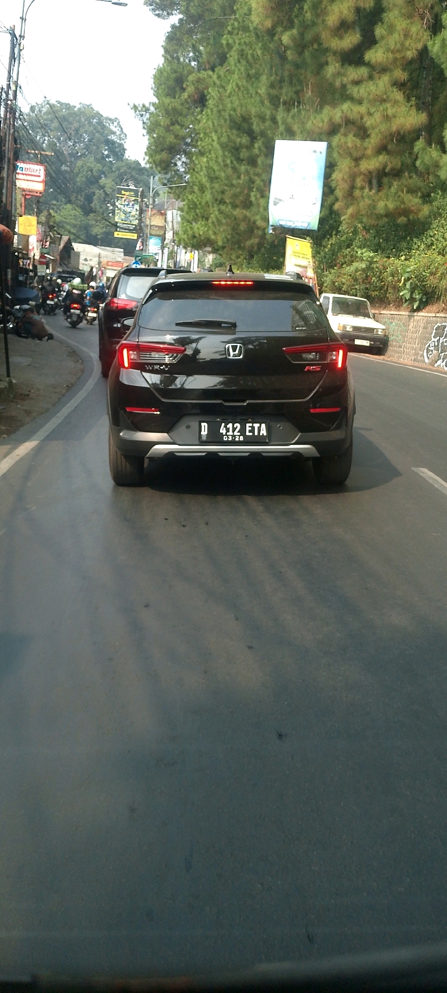 The image captures a rear view of a black Honda WR-V car with license plate D 412 ETA, registered in Bandung, Indonesia. The car is in motion on a paved road, likely a street in Bandung, given the license plate format. In the background, there is a red car and several motorcycles, indicating traffic. The environment features tall trees, suggesting a location with significant greenery, possibly a suburban or hilly area of Bandung. Advertising banners are visible on poles along the roadside, typical of urban streetscapes. The sky is overcast, casting diffused light on the scene. The overall impression is of a typical day in traffic within the city of Bandung.