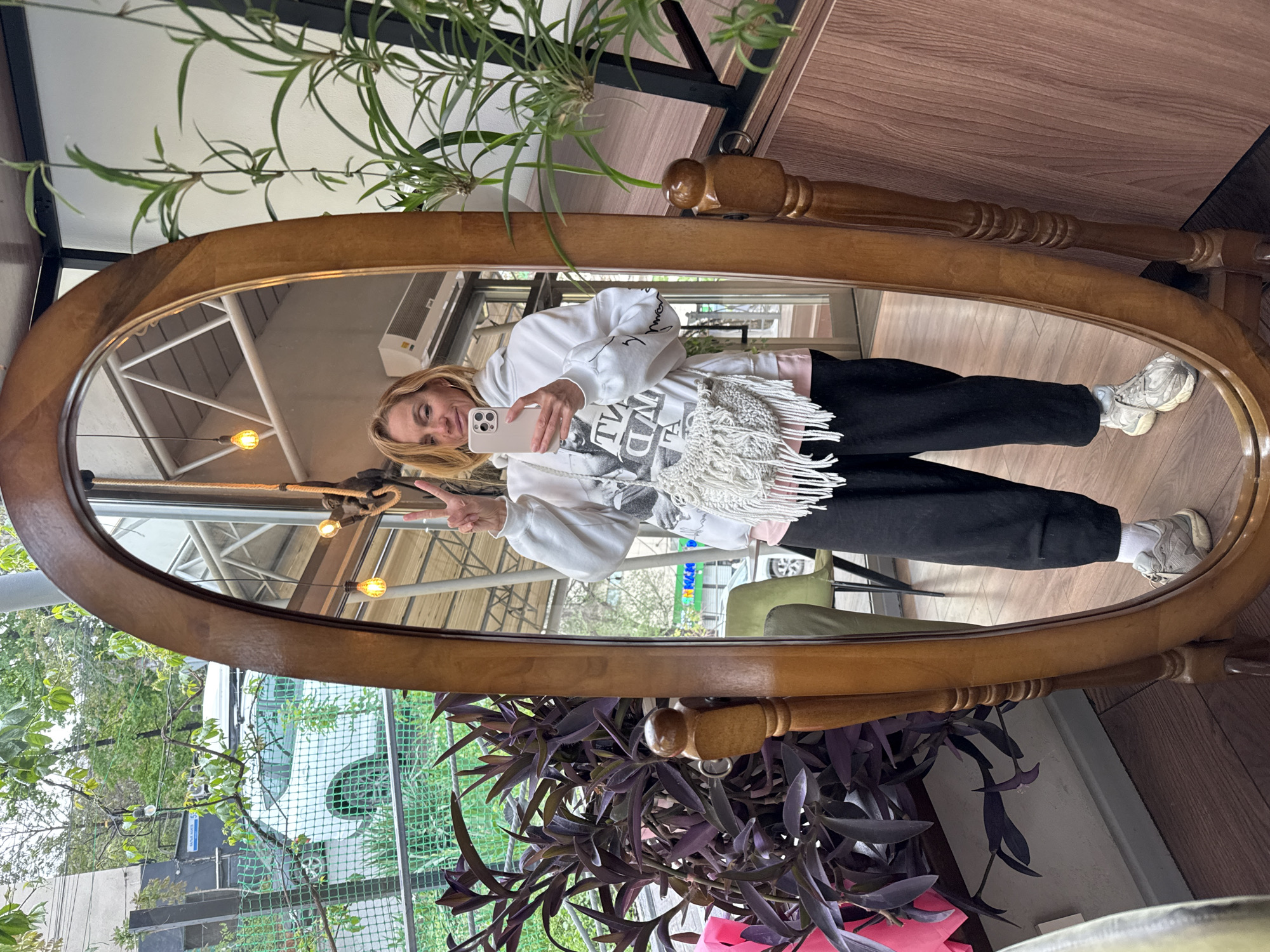 The image displays a mirror selfie of a woman taken indoors, likely within a cafe or well-appointed establishment in Bishkek City, Kyrgyzstan. The woman, with blonde hair, is reflected in a large, oval, wooden-framed mirror, holding a light-colored smartphone to take the picture and making a peace sign with her left hand.
She wears a white hooded sweatshirt featuring black graphics and partially visible inverted text that reads "LIVE TRUTH" on the front, and additional text on the sleeve. A white, fringed accessory is draped over her shoulder. She is also wearing black pants and light-colored sneakers with white socks.
The interior reflected in the mirror shows an exposed ceiling structure with metal beams, a white air conditioning unit, and warm yellow pendant lights. Surrounding the mirror and within the reflection are various potted plants, including vibrant green leafy plants and others with deep purple foliage, contributing to a lively atmosphere.
Through a window reflected in the mirror, an outdoor urban scene is visible, featuring a white vehicle, a green mesh fence, and more trees and buildings. The lighting suggests it is daytime, possibly overcast. The woman's expression is friendly and casual.