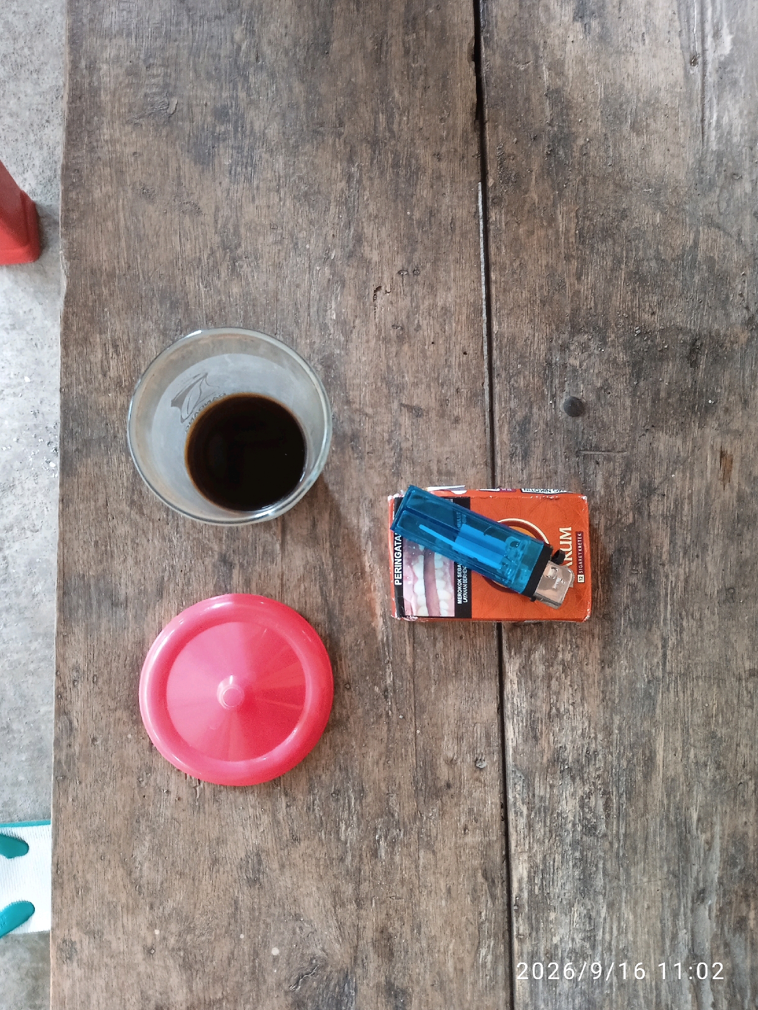 A close-up, overhead shot shows a collection of items on a weathered wooden surface, possibly a table. In the upper left quadrant is a clear glass containing a dark liquid, presumably coffee. Below the glass, a bright red circular lid is placed on the wood. To the right of the lid is a pack of cigarettes, predominantly orange with text in Indonesian, and a blue plastic lighter resting on top. The wooden surface exhibits visible grain and some wear. The timestamp "2026/9/16 11:02" is visible in the lower right corner, suggesting the image was captured in Kelurahan, Indonesia.