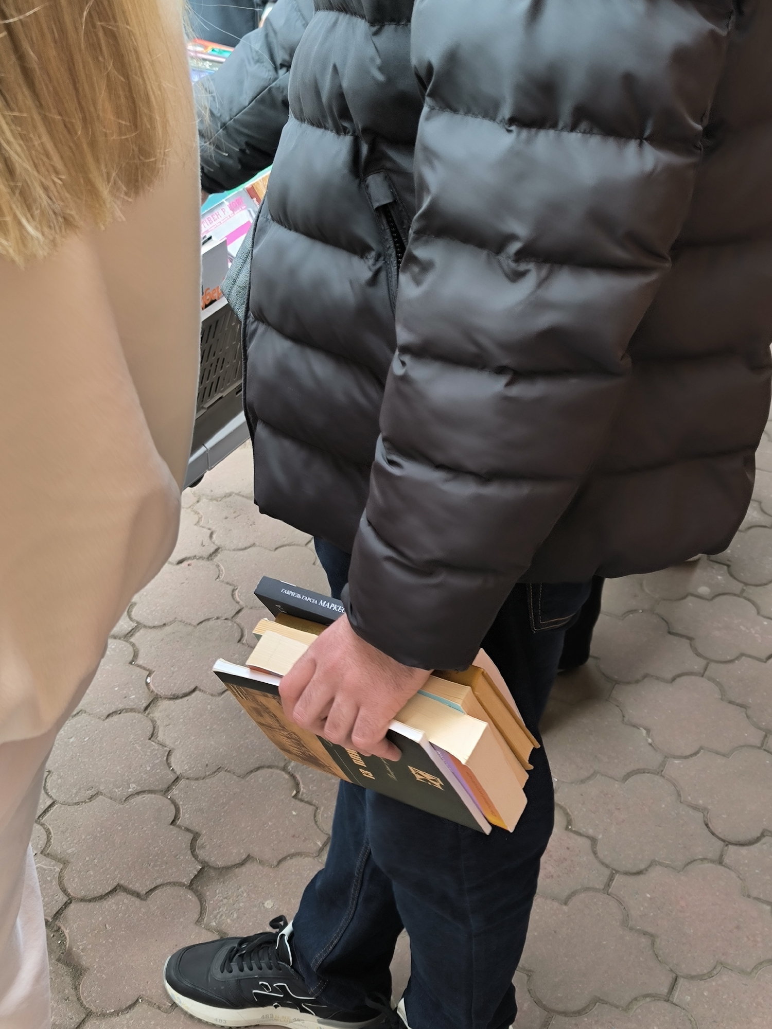 A medium shot depicts two individuals standing on a brick-paved surface. To the left, a portion of a person with blonde hair is visible. To the right, an individual clad in a dark, puffy jacket and dark denim jeans holds a stack of books. One of the books appears to have the title "Gabriel Garcia Marquez" on its spine. The person is also wearing black sneakers with white accents. The brick paving is arranged in a repeating cloud-like pattern. The location is Vinnytsia, Ukraine.