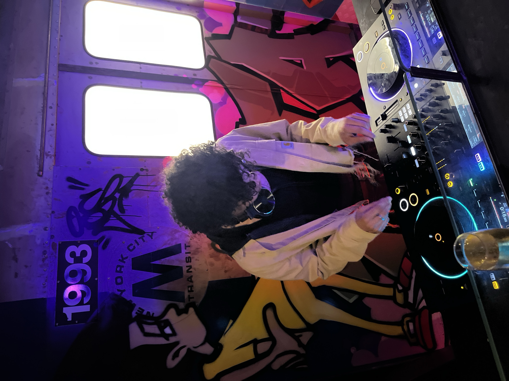 A DJ with curly hair is operating a DJ controller at a Barcelona venue. The DJ is wearing a light-colored jacket over a dark shirt, and headphones are around their neck. The equipment is lit with blue and white lights. A partially visible glass appears to be on the DJ table.
The wall behind the DJ is decorated to resemble a New York City subway car, with graffiti and stylized imagery, including the text "1993 NEW YORK CITY TRANSIT". Two illuminated windows are visible at the top of the frame.