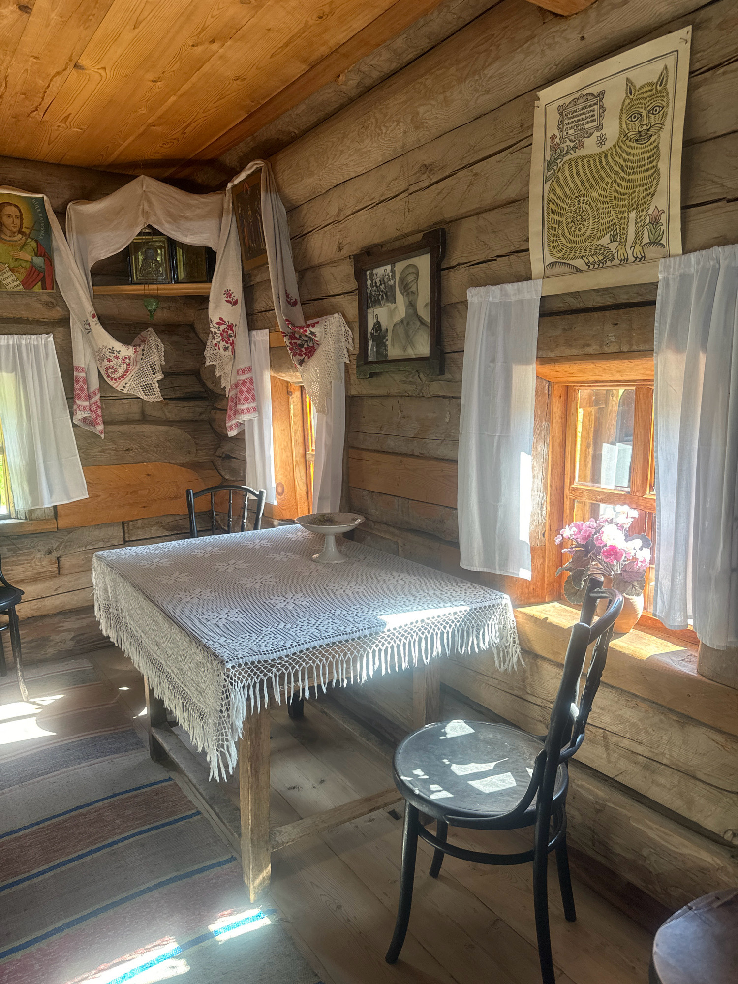 The image depicts the interior of a traditional wooden cabin, likely a museum exhibit, within the Taltsy Museum of Wooden Architecture and Ethnography in Irkutsk, Russia. Sunlight streams through two windows, illuminating a rustic setting. In the center, a wooden table is draped with a white, lacy tablecloth. On the table sits a decorative pedestal with a shallow bowl. Two dark wooden chairs, one with a bentwood design, are positioned near the table.
The cabin walls are constructed from rough-hewn logs. The ceiling is also made of wooden planks. Various decorative and religious items adorn the walls and shelves. To the left, an icon of a saint is visible. Above the table, a white cloth with embroidered red and pink floral patterns hangs, draped to create a canopy over icons and other religious artifacts on a shelf. To the right, a folk art print of a stylized cat with text in a frame is mounted on the wall. Below it, a framed black and white photograph of men in military-style uniforms is displayed.
White curtains hang from the window frames, softening the sunlight. A vase filled with pink flowers sits on the windowsill of the right window. A striped rug with bands of red, blue, and brown lies on the wooden floor, its pattern partially visible beneath the table and chair. The overall impression is one of a preserved historical domestic interior, showcasing traditional Russian rural life and possibly religious practices.