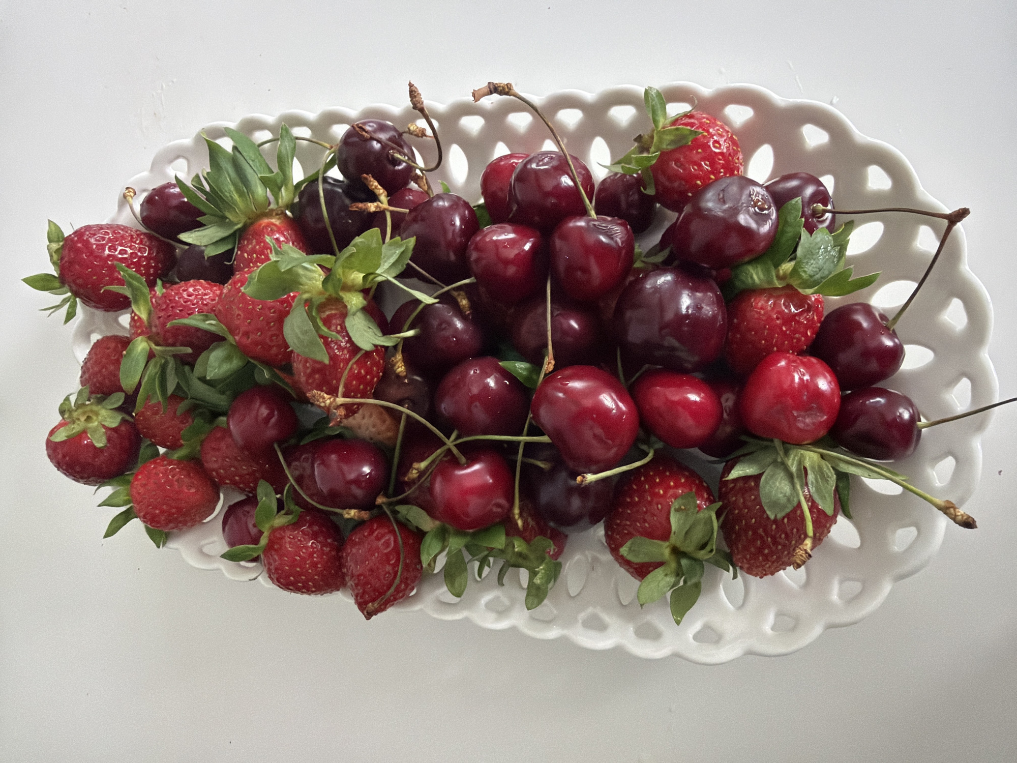 A close-up, top-down view of a white decorative platter filled with fresh strawberries and cherries. The platter is oval and has an intricate, lace-like edge with cut-out patterns. The fruits are artfully arranged, with the bright red of the strawberries and the deep red of the cherries creating a vibrant contrast. Green leaves are visible amongst the fruit, adding to their freshness. The fruits are predominantly clustered in the center of the platter, spilling slightly towards the edges. The background is a plain white surface, likely a table, which makes the colorful fruits stand out. The lighting is bright and even, suggesting an indoor setting with good illumination. There are no people visible in the image, and no specific actions are depicted beyond the presentation of the fruit. The scene is static and focused solely on the arrangement of the fruit on the platter. The location in Baloži, Latvia, is not visually indicated by any landmarks or environmental cues in the image.