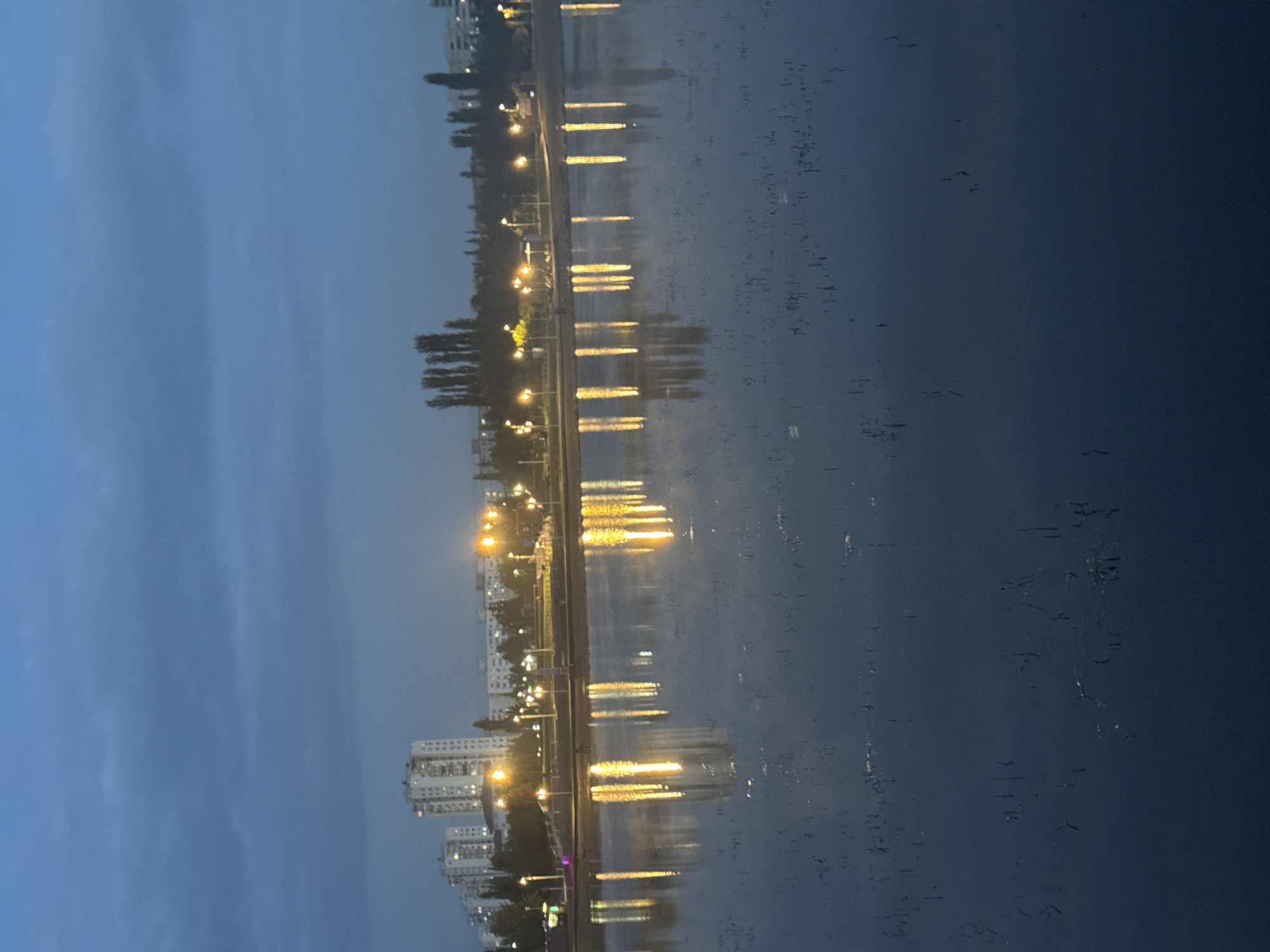 The image depicts an evening scene in Ufa, Russia, likely at the Ufa Amphitheater or a similar waterfront park area. The sky is a deep twilight blue with a hint of clouds. In the background, several multi-story apartment buildings are visible, illuminated by interior lights. A row of streetlights line a pathway along the waterfront, casting a warm, yellowish glow. These lights are reflected in the calm water of a lake or river, creating elongated, shimmering streaks of light. The water's surface is disturbed by what appear to be small, dark reeds or aquatic plants, breaking up the reflections in places. The overall atmosphere is serene and tranquil, characteristic of an urban park at dusk. There are no people or animals clearly visible in the scene.