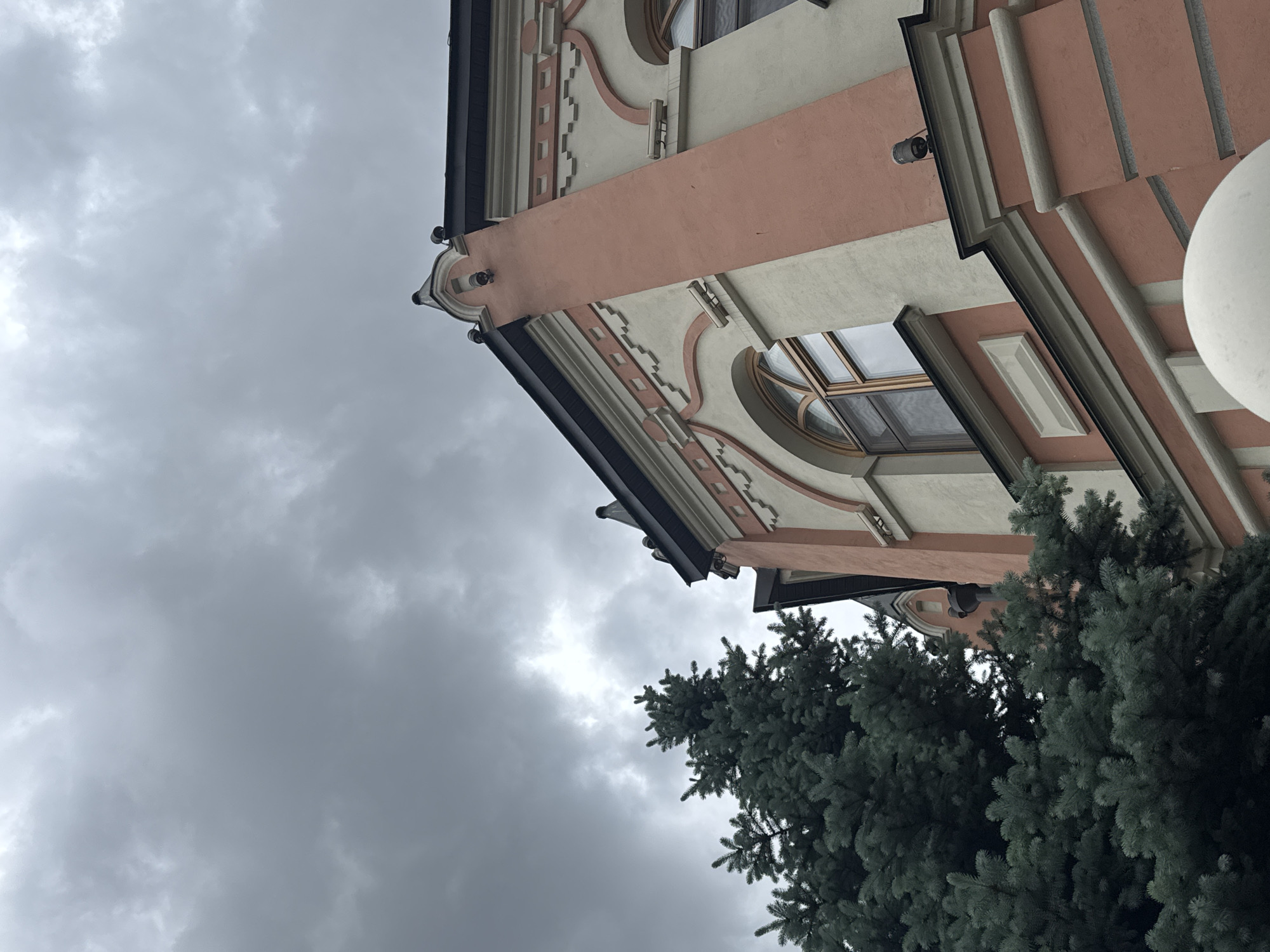 The image shows a building facade in Moscow, Russia, under a cloudy sky. The building is multi-storied and features light-colored walls with peach-colored accents. There are arched windows on the facade. An evergreen tree partially obscures the lower part of the building on the right side. The photograph is taken from a low angle, looking upwards towards the building.