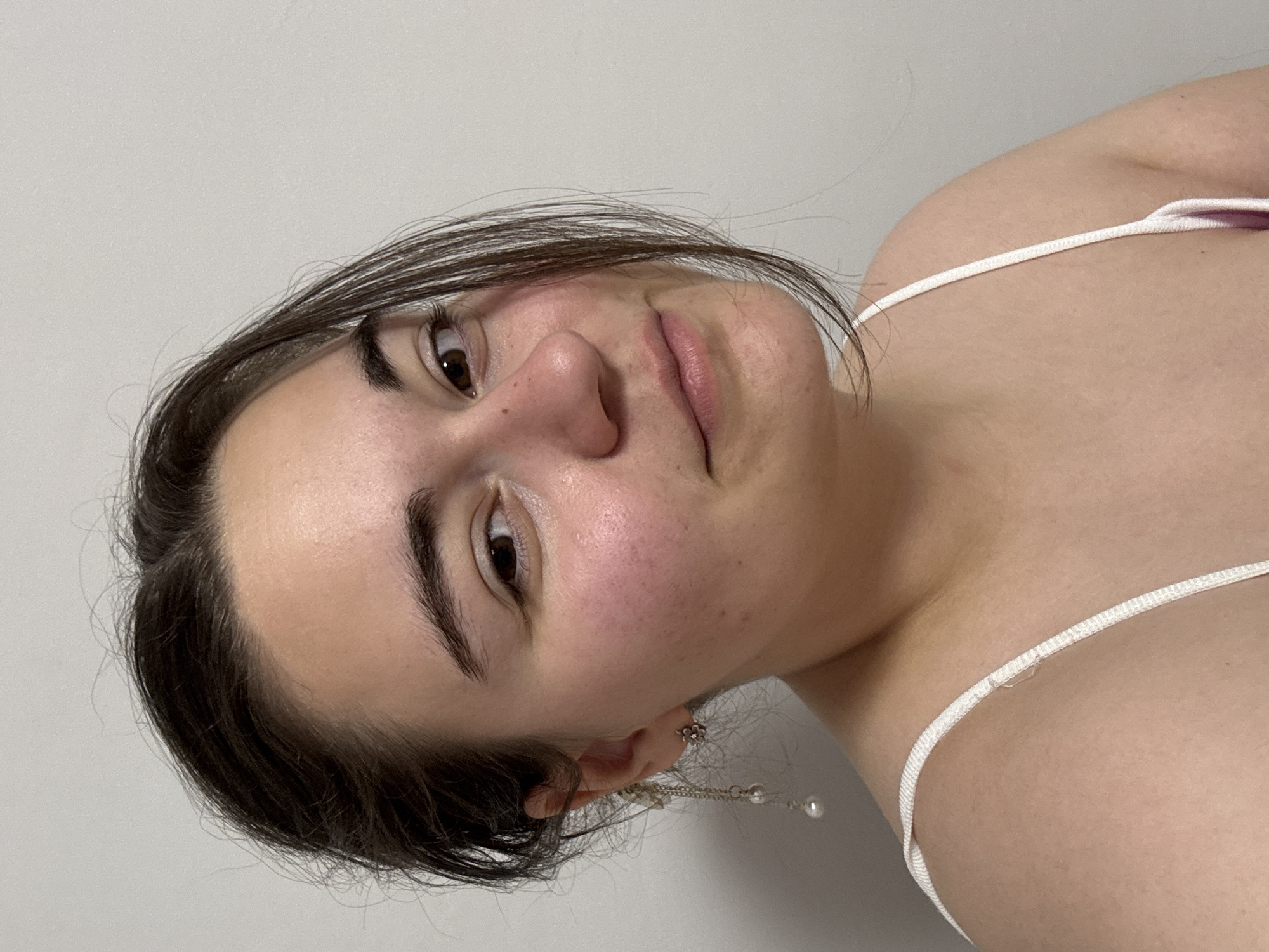 A close-up portrait of a young woman with dark hair. She has brown eyes, full eyebrows, and fair skin. She is wearing a white tank top with thin straps and dangling earrings. The background appears to be a plain white wall. The image does not provide information about the location or any specific actions taking place. There is no indication the location is Zlatitsa, Bulgaria.