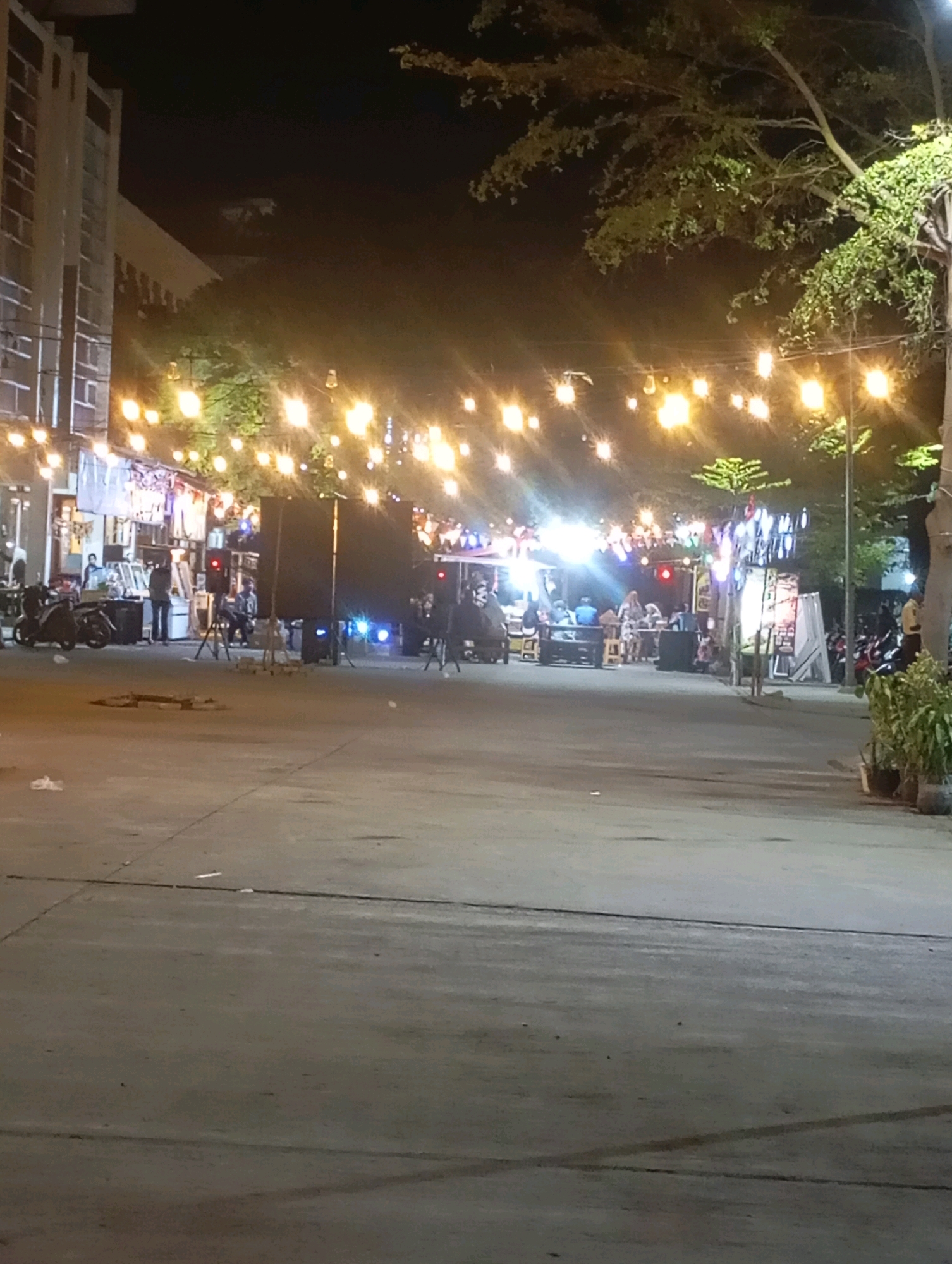 An outdoor street scene at night in Babelan, Indonesia, features a row of shops or stalls illuminated by strings of festive lights. A paved street runs through the foreground, leading to the stalls in the background. Several motorcycles are parked along the left side of the street. In the middle ground, large speaker systems are set up, suggesting a possible event or performance. People are visible in small groups, some seated at tables outside the stalls, and others standing. The environment is dimly lit, with the primary light sources being the string lights and the illuminated interiors of the stalls. Trees with foliage are present along the right side of the scene, casting shadows. The overall impression is of a lively night market or street fair.