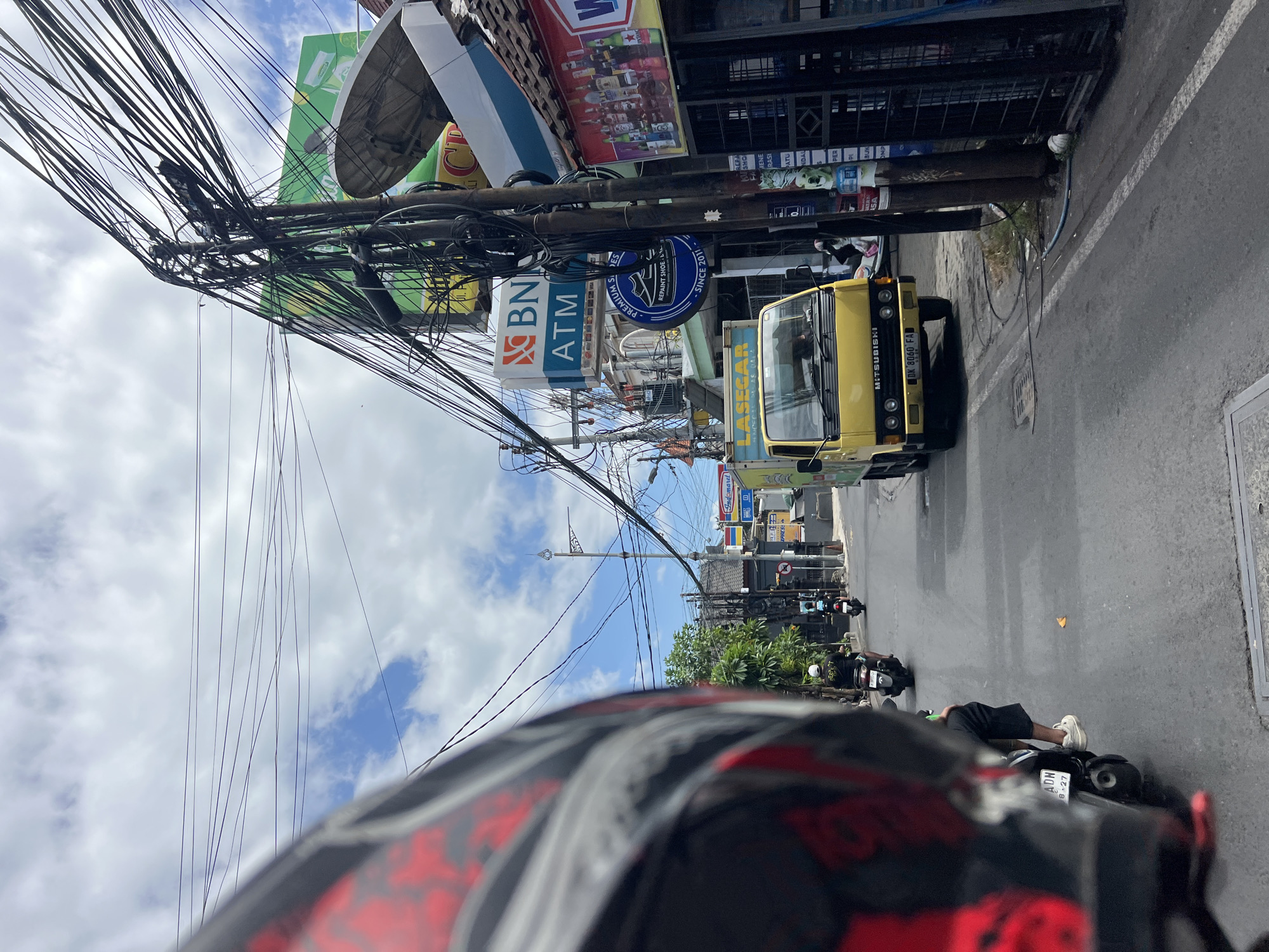 In Kuta, Indonesia, a street scene unfolds with a prominent motorcycle helmet in the foreground, obscuring the rider's perspective of the bustling road. A bright yellow Mitsubishi truck, bearing license plate "DK 8060 FA," is captured mid-travel, heading away from the viewer. The road is lined with various businesses and signs, including "BNI ATM," "Indomaret," and advertisements for "Premium Shoes Since 2011." Numerous electrical wires crisscross the sky, strung between utility poles, adding to the urban density. Several other motorcycles are visible on the road, with riders navigating the traffic. The sky above is partly cloudy, with a mix of blue sky and white clouds. The overall environment suggests a typical day in a busy commercial district of Kuta.