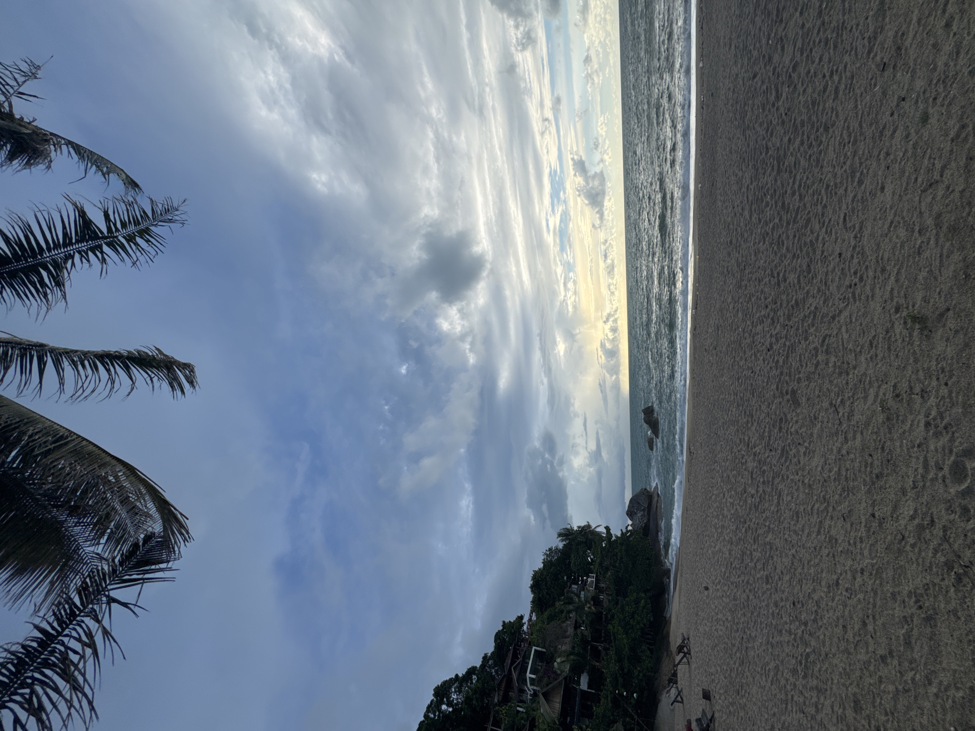 The image depicts a beach scene in Baan Thong Nang, Thailand. The foreground is dominated by sandy beach. The shoreline is visible with waves. Palm trees are present on the left side and buildings are embedded in lush vegetation on a hill near the coast. The sky is overcast with layered clouds. There are people in the distance.
