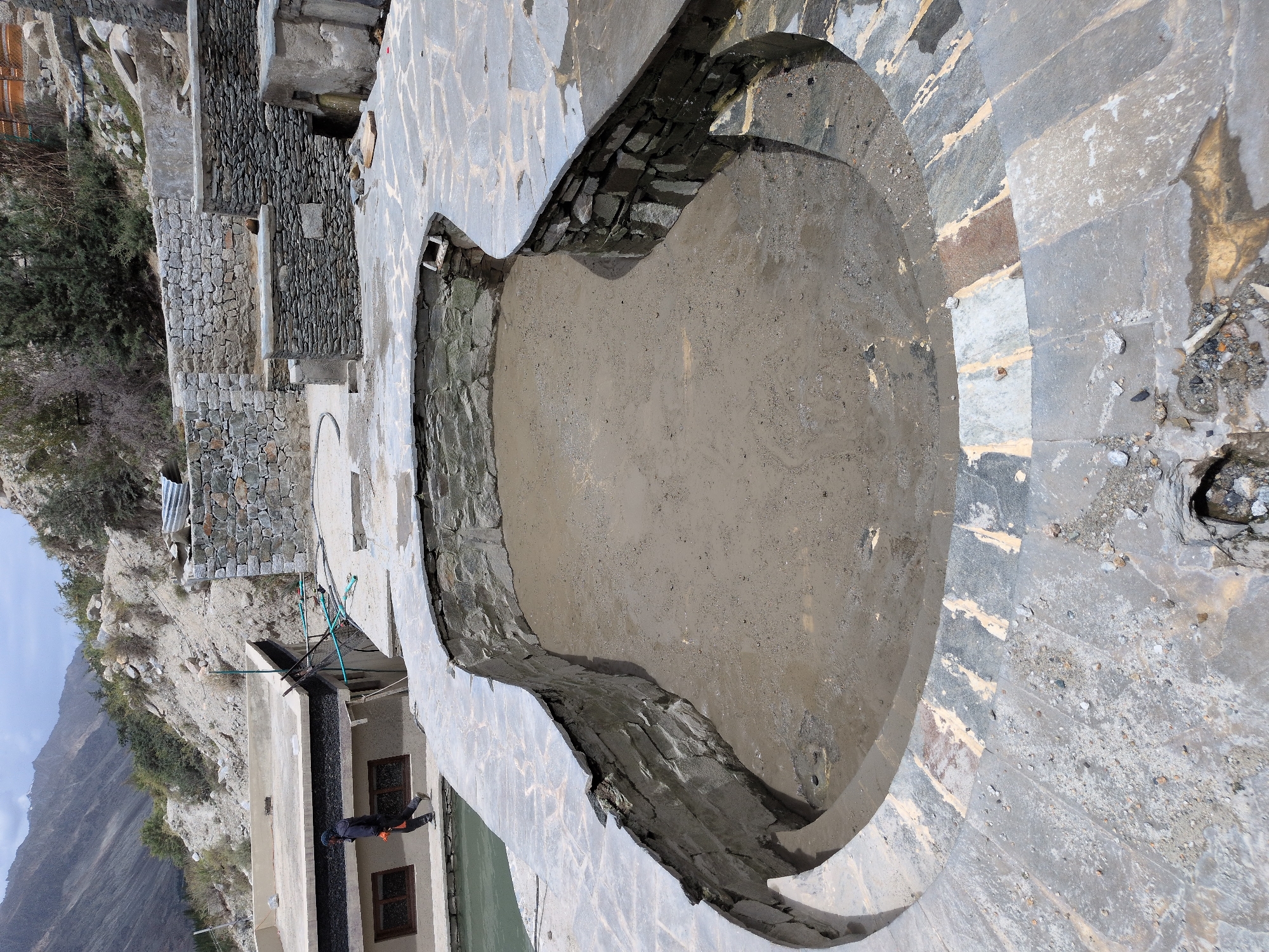 The image depicts an outdoor scene in the city of Panamik, India, focusing on a partially constructed or renovated outdoor pool area. The pool itself is empty and filled with a muddy, sandy substance, with its stone-lined walls visible. The surrounding area is paved with irregular, flat stone tiles, some of which appear to be in the process of being laid or repaired, with exposed mortar and debris.
In the background, a traditional building with a flat roof and windows is visible, and a person dressed in dark clothing with an orange tool in hand is walking along the edge of the pool area. Further in the background, a rocky, mountainous landscape under a cloudy sky suggests a rural or mountainous setting. Stone walls and structures, some appearing aged and others more recently built with smaller stones, are integrated into the hillside, characteristic of the region's architecture. The overall impression is one of ongoing construction or maintenance in a remote, natural environment.