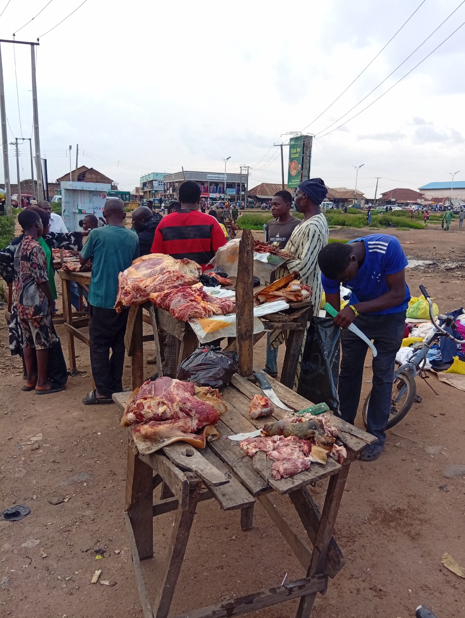 This is a photograph of an outdoor meat market in Bukuru, Nigeria. The scene is set on a dusty, unpaved ground with several wooden tables serving as makeshift butcher stalls. A cloudy sky suggests it might be late afternoon or overcast.
Several people are present, appearing to be vendors and customers. In the foreground, a young man in a blue t-shirt is actively cutting meat with a long, curved knife. He is focused on his task, with a bicycle and bundles of clothing to his right. On the tables, large cuts of raw meat are displayed, some appearing to be beef or pork. Several knives are visible on the tables, indicating ongoing butchering and preparation.
Other individuals are seen in the mid-ground and background. Some appear to be interacting with the vendors, while others are standing around. The overall atmosphere seems bustling and typical of a local market. In the distance, there are buildings, some appearing to be shops or stalls, and more people can be seen. A large green billboard with text that includes "Eat for Good" is visible in the upper right portion of the image, providing a clear location cue. Power lines are strung across the sky, a common sight in many urban and semi-urban areas in Nigeria.