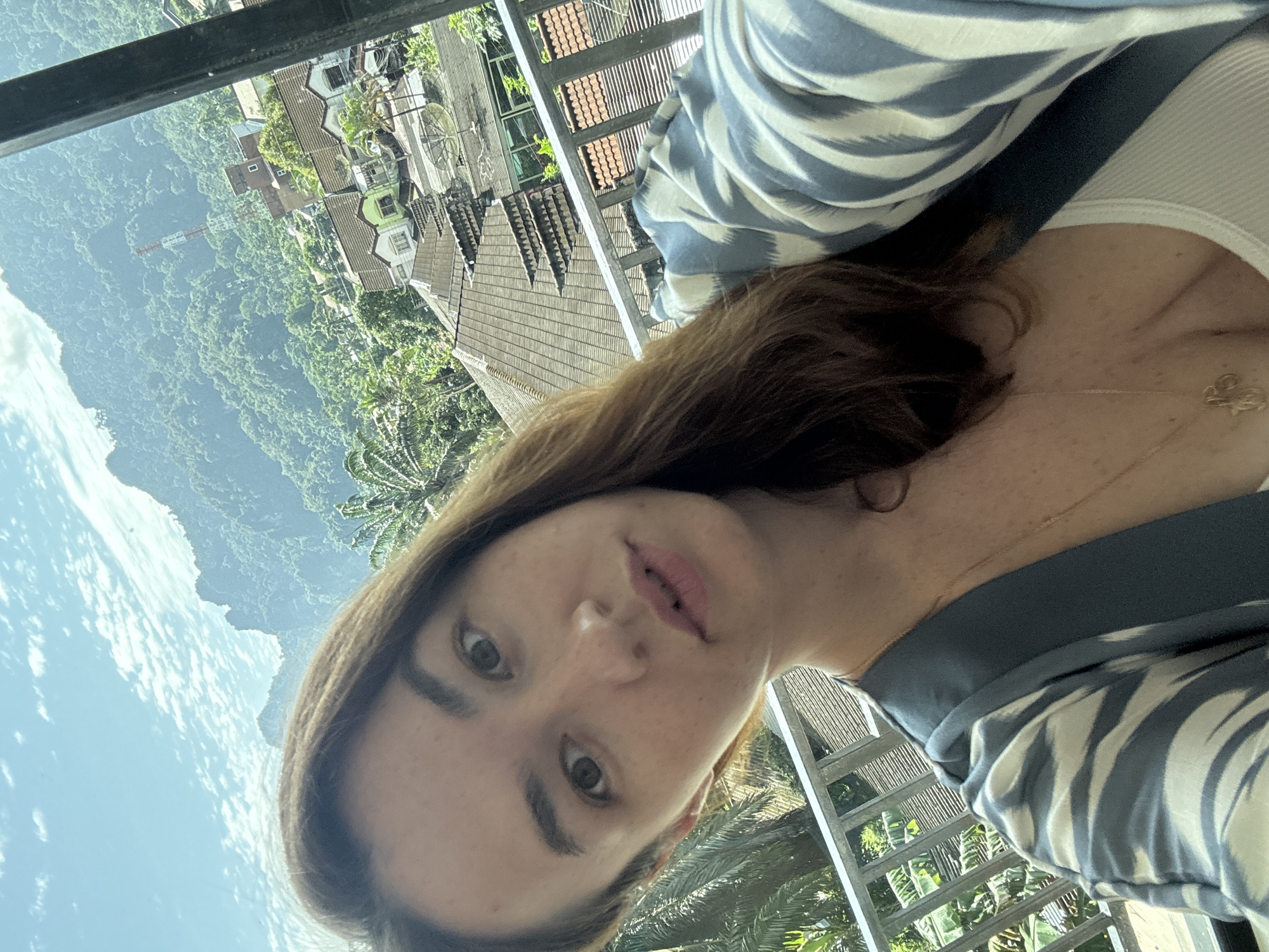 A woman is taking a selfie from a balcony overlooking a town and mountains in Phang-nga, Thailand. She has long brown hair and is wearing a blue and white striped robe over a white tank top. The background shows lush green mountains, several buildings with tiled roofs, and a communication tower. The sky is blue with scattered clouds.