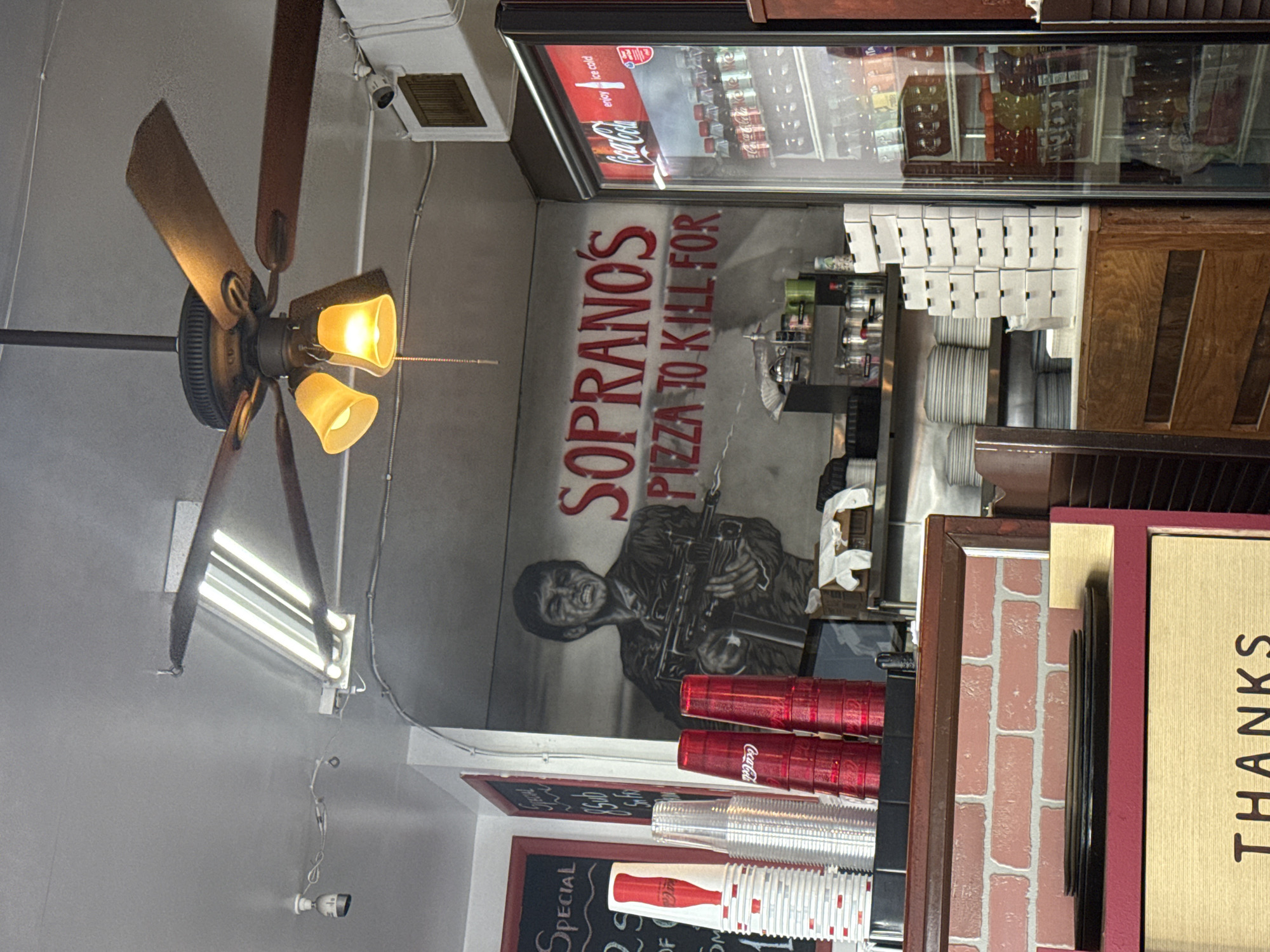 The image captures the interior of Soprano's Pizza in Ocean City, United States of America. A vintage ceiling fan with three blades and illuminated light fixtures hangs prominently from the ceiling. A mural on the wall displays the words "Soprano's Pizza to Kill For" above a stylized depiction of a gangster holding a machine gun. A glass-front beverage cooler is visible in the background, stocked with drinks. A stack of red and white plastic cups are stacked on a shelf, while a chalkboard menu with the word "Special" is partially visible. A "Thanks" sign in gold lettering is positioned in the lower right corner. The scene is somewhat cramped, with various kitchen and service-related items visible around the edges of the frame.