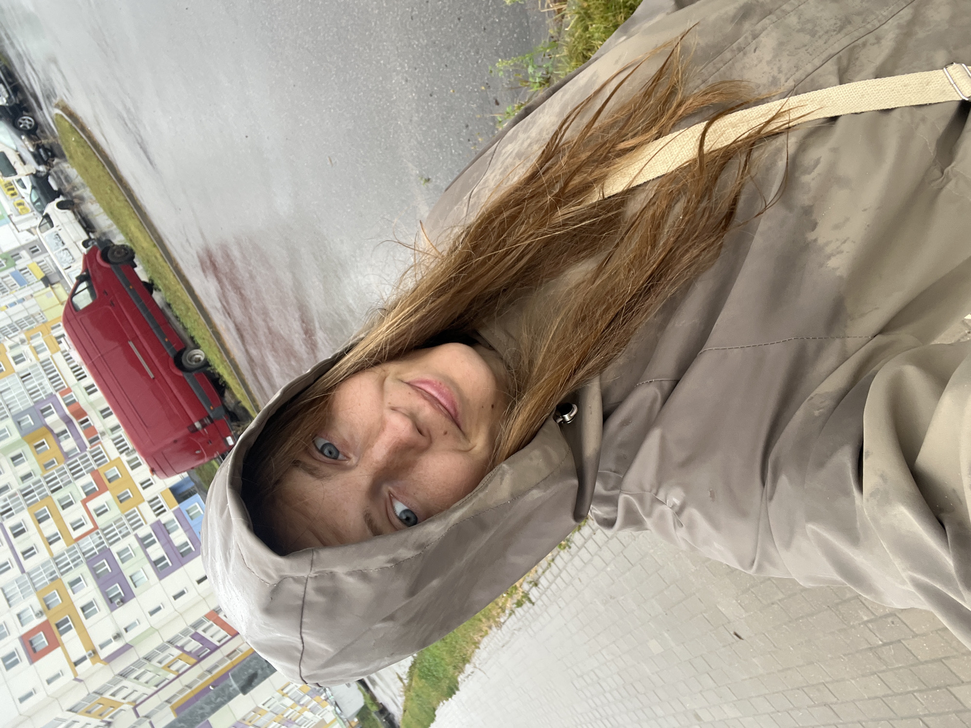 A woman with long, wet, light brown hair is taking a selfie outdoors on a paved walkway next to a street. She is wearing a beige hooded raincoat, and her face is visible, looking at the camera with blue eyes and a slight smile. Her hair is damp, and there are water droplets on her raincoat. In the background, a colorful apartment building with many windows is visible, along with a red van and several other parked vehicles on the wet street. The ground is also wet, indicating it is raining. The location is identified as Senitski Sel'ski Sovet, Belarus.