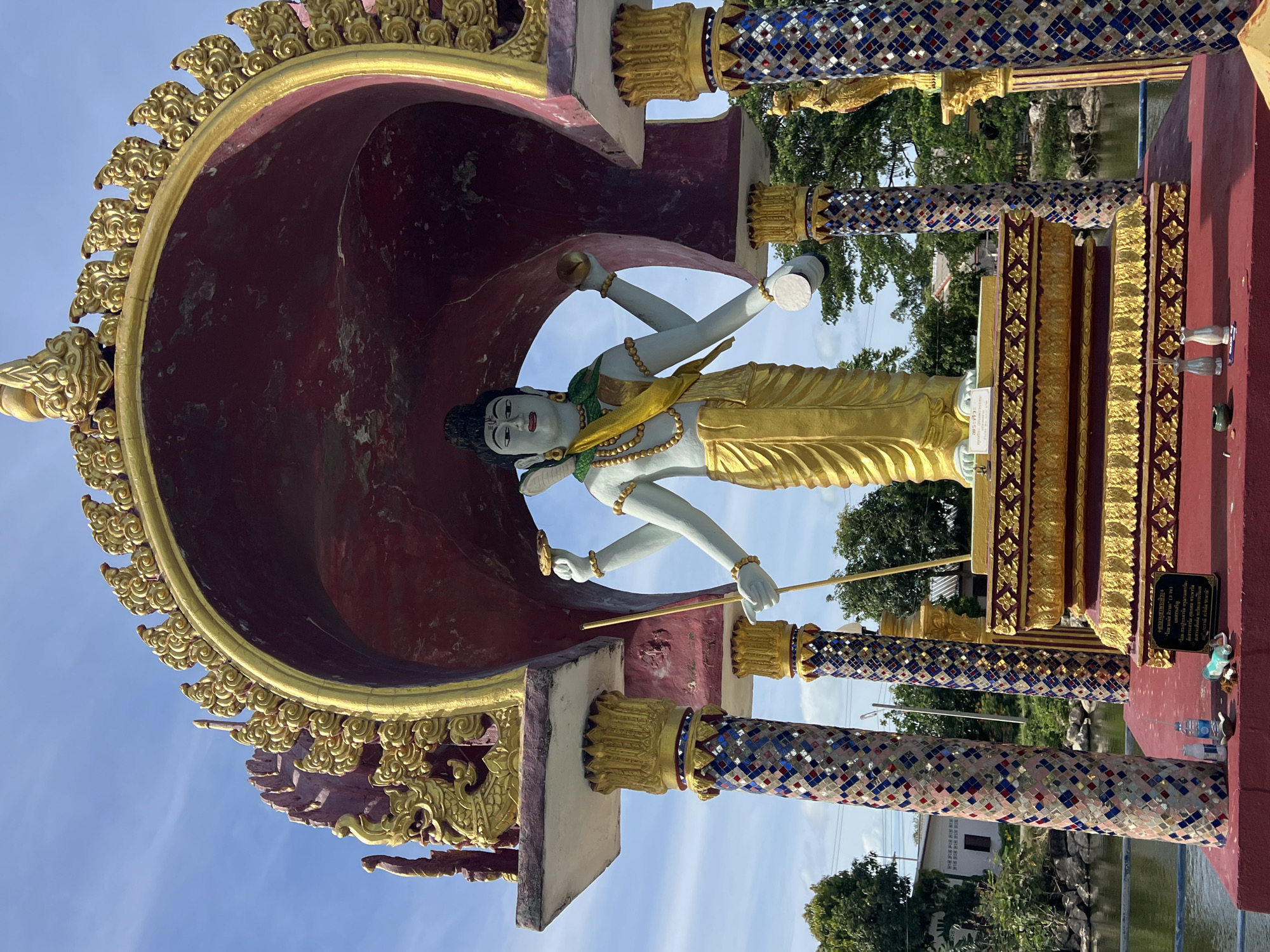 A statue of the Hindu deity Shiva, depicted with multiple arms and a serene expression, stands within a vibrantly decorated architectural structure. The statue is light blue-skinned, adorned with golden garments and jewelry, and wears a green sash and a yellow cloth draped over its shoulder. In its hands, Shiva holds various symbolic objects, including a staff, a drum, and what appears to be a disc.
The statue is positioned on a raised, ornate golden platform, which itself rests on a red base. The structure enclosing Shiva features ornate golden flourishes and is painted a deep red on the interior. Four decorative columns support the overhead structure. These columns are cylindrical and decorated with a mosaic of small, colorful tiles in shades of blue, red, and white, with golden capitals.
The scene is set outdoors, with a bright blue sky and scattered white clouds visible in the background. Lush green trees and foliage surround the structure. In the distance, a body of water, possibly a pond or canal, is visible, reflecting the sky. A small building with a white facade and a dark roof can be seen to the left, behind the water.
On the red base in front of the statue, various offerings are present, including small bottles, a water bottle, an incense holder with burnt incense sticks, and what appears to be a small, white offering. A plaque with Thai script is also visible on the base. The overall impression is one of a religious or cultural site, likely a temple or shrine, located in a tropical environment. Based on the architectural style and the presence of Hindu iconography, this location is likely in Thailand, and given the context of Ko Samui, it is plausible this is a shrine within that district.
