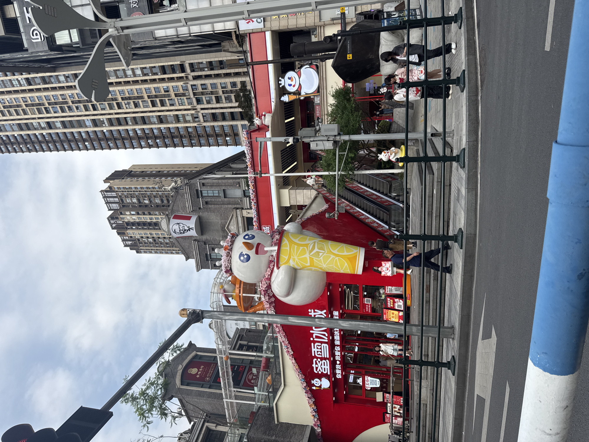 The image captures a street scene in Nan'an District, China. The architecture is a mix of modern high-rise buildings and traditional-style structures. A prominent feature is a large, cartoonish snowman figure holding a cup of lemon drink, likely a promotional display for a 雪冰成 (Mì Xuě Bīng Chéng) store, a chain known for its ice cream and tea. The store is painted in red with white lettering and decorations. An escalator can be seen in the background. Pedestrians are walking along the sidewalk. Street furniture, including lamp posts and barriers, are visible. A KFC restaurant is partially visible in the upper portion of the image. The sky is overcast.