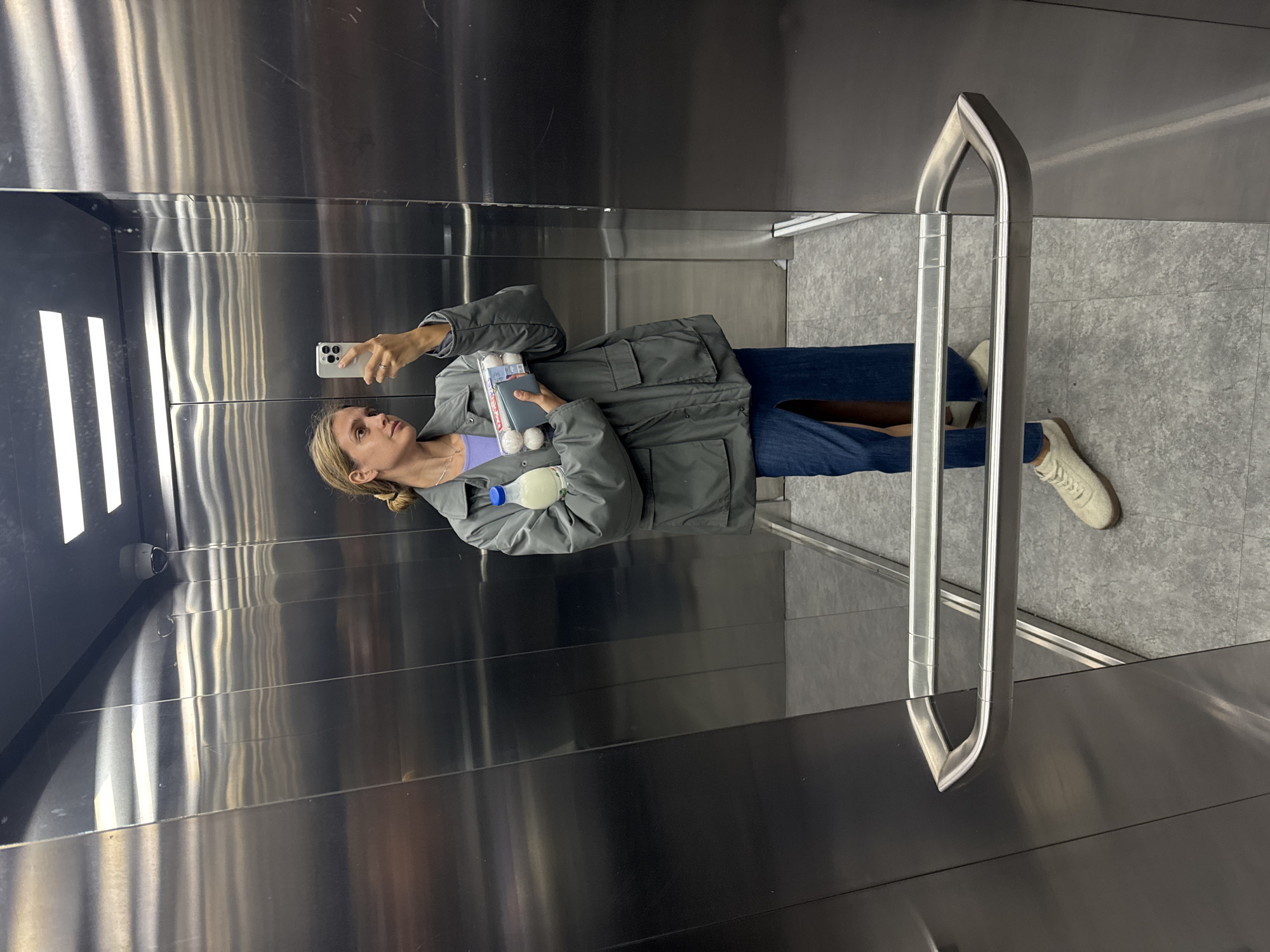 A young woman is depicted taking a selfie inside a modern elevator with stainless steel walls and ceiling. The image is oriented sideways, with the woman appearing to lie horizontally against the reflections of the elevator interior.
The woman has fair skin and light brown hair tied back, with some strands visible around her face. She is wearing a light purple top, a practical grey-green jacket with large pockets, blue jeans, and light-colored sneakers. In her right hand, she holds a white/silver smartphone, angled as if capturing her reflection in the elevator wall. Her expression is neutral, focusing on the phone.
Under her left arm, she carries several items. Visible are two small, white, spherical or ovate objects, possibly eggs or similar packaged goods, and a small, dark rectangular object, which could be a wallet or cardholder. Additionally, a clear plastic bottle containing a white liquid (likely milk or a dairy drink) with a blue cap is tucked securely under her arm.
The elevator interior is characterized by highly reflective, brushed stainless steel panels that show distorted reflections of the woman, the handrail, and interior lights. Vertical strip lights are visible on one side wall, casting bright reflections across the metallic surfaces. The floor is covered with grey tiles. A metal handrail runs along one side of the elevator. In one corner, near the ceiling, a small security camera is visible. No discernible text is clearly legible on any of the objects or surfaces. The scene is brightly lit by artificial light sources within the elevator.