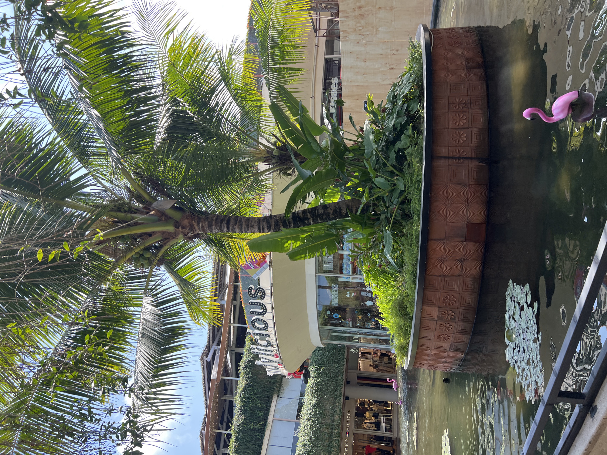 A wide shot captures a serene water feature in Kuta, Indonesia, likely within a shopping complex, featuring lush greenery and a prominent palm tree. The water, reflecting the bright sky and surrounding architecture, holds lily pads and two pink flamingo statues. One flamingo is closer to the foreground, while another is further back near a store with a "Tommy Hilfiger" sign. The background shows multi-level buildings covered in greenery and a store with a sign reading "Mouth Candy," and another possibly named "Hersheys." A dark railing in the foreground suggests a viewing area or walkway. The scene is bathed in sunlight, indicating a clear day.