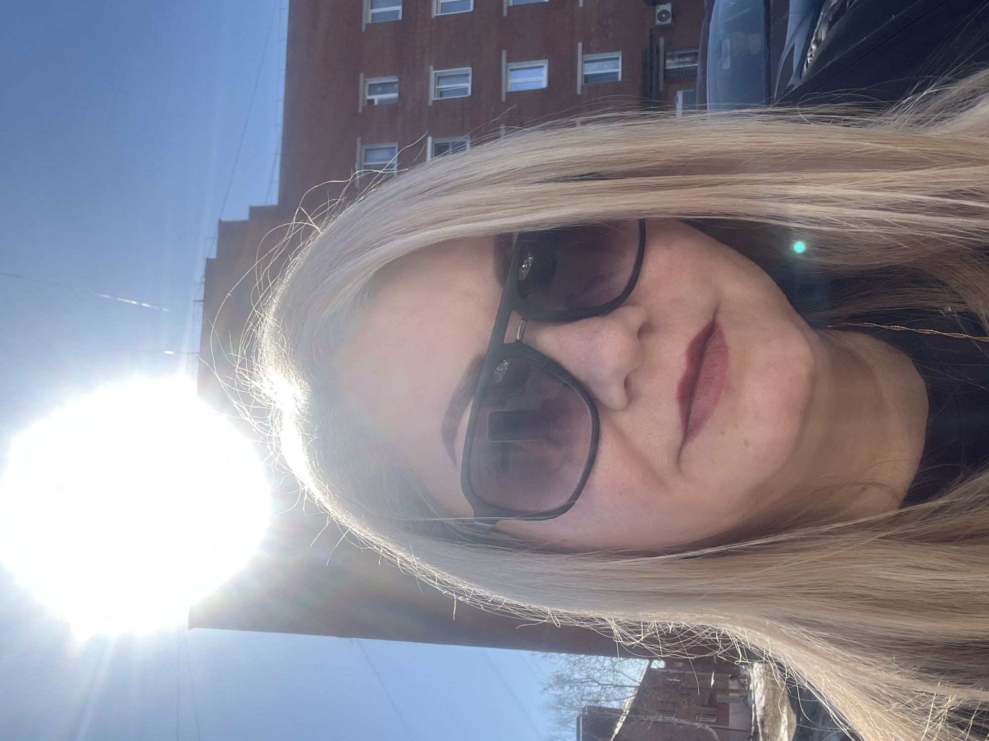 A woman with blonde hair is taking a selfie outdoors in the city of Glazov, Russia. She is wearing sunglasses and has red lipstick on. Behind her is a red brick building with many windows. The sky is blue and sunny, with the sun's glare visible in the bottom left corner of the image. A car is partially visible in the top right corner.