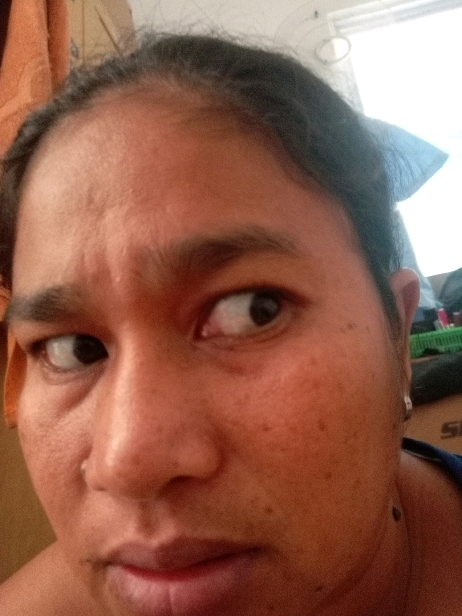 A close-up, over-the-shoulder shot shows the left side of a person's face, with their eyes looking to the left. The person appears to be in an indoor setting with a neutral background. The lighting suggests it is daytime, possibly near a window, as indicated by the bright light on the right side of the image. The person's skin tone is medium brown, and their features are clearly visible, including their eyebrows, eyes, nose, and mouth. There is some texture on the skin, with small pores and a few moles visible. The person's hair is dark and appears to be pulled back. The overall impression is a candid, personal photograph taken in a domestic environment. The provided information about Batam, Indonesia, is not visually represented in the image itself.