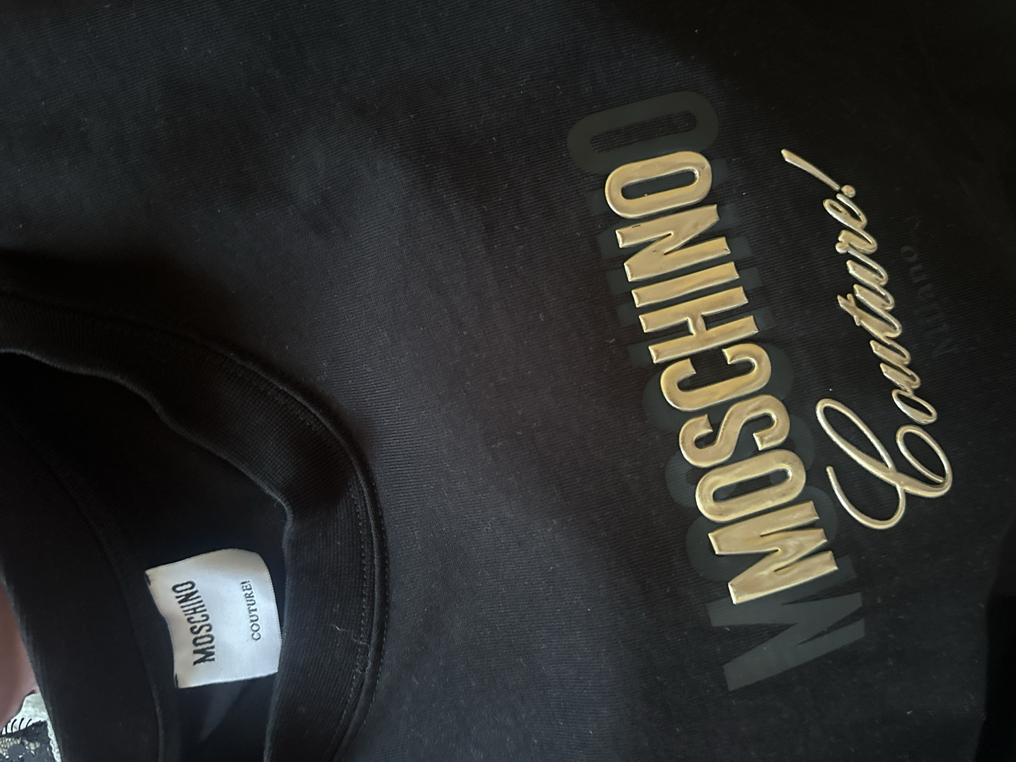 The image shows a close-up of a black t-shirt with "MOSCHINO" and "COUTURE!" printed on it. The t-shirt is made of cotton and has a ribbed neckline. The print is in a metallic gold color. The t-shirt is lying flat on a surface, which appears to be another piece of fabric. There is a label on the inside of the t-shirt that also reads "MOSCHINO COUTURE!". The background is dark and out of focus, making it difficult to determine the environment.