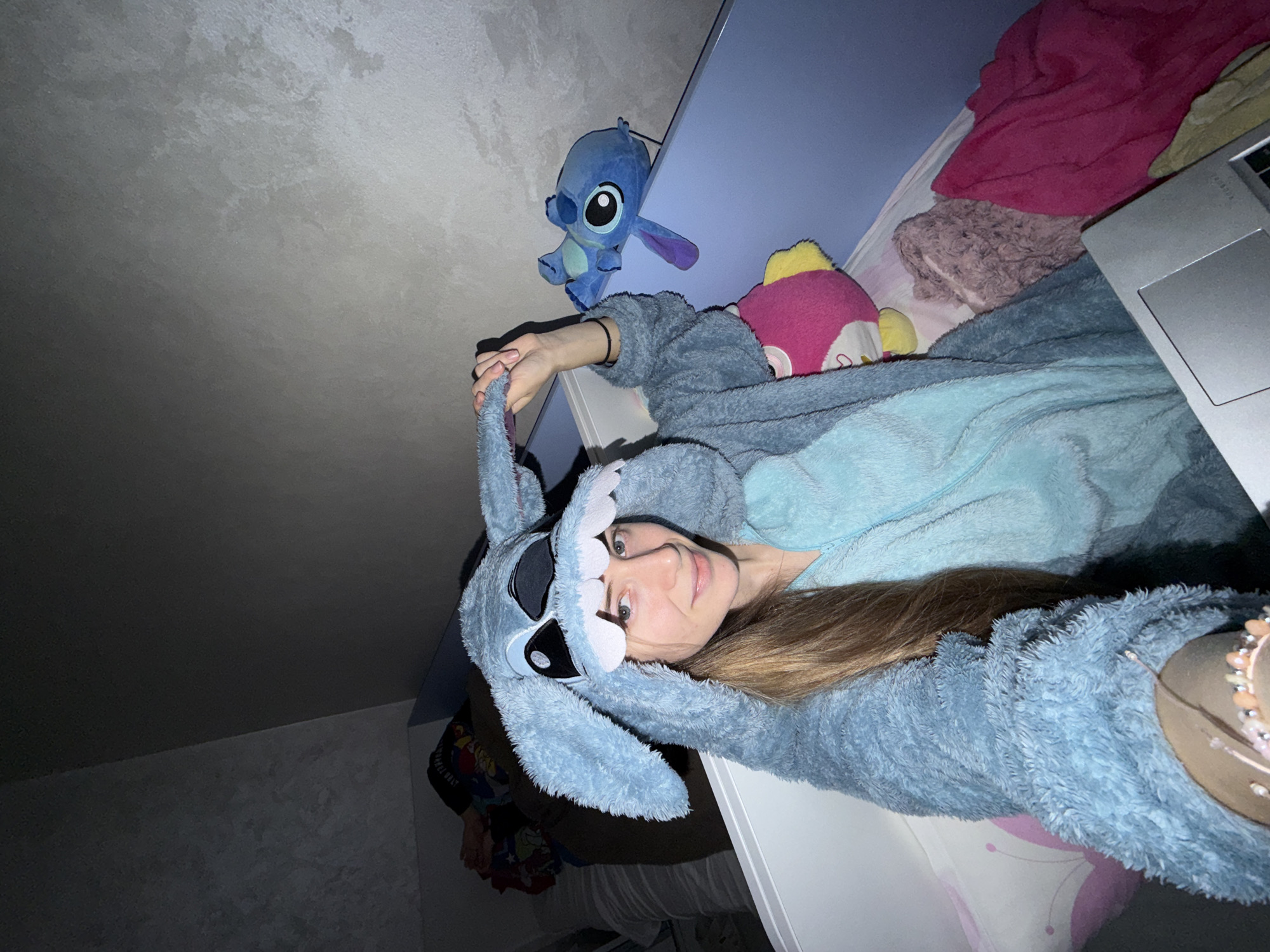 A person wearing a blue Stitch onesie is lying in bed, taking a selfie. A blue Stitch plush toy is visible on the bedpost. The room appears to be a bedroom, possibly in a home, and there is a bed with various blankets and pillows. There are no clear indications of the location being Plovdiv, Bulgaria.