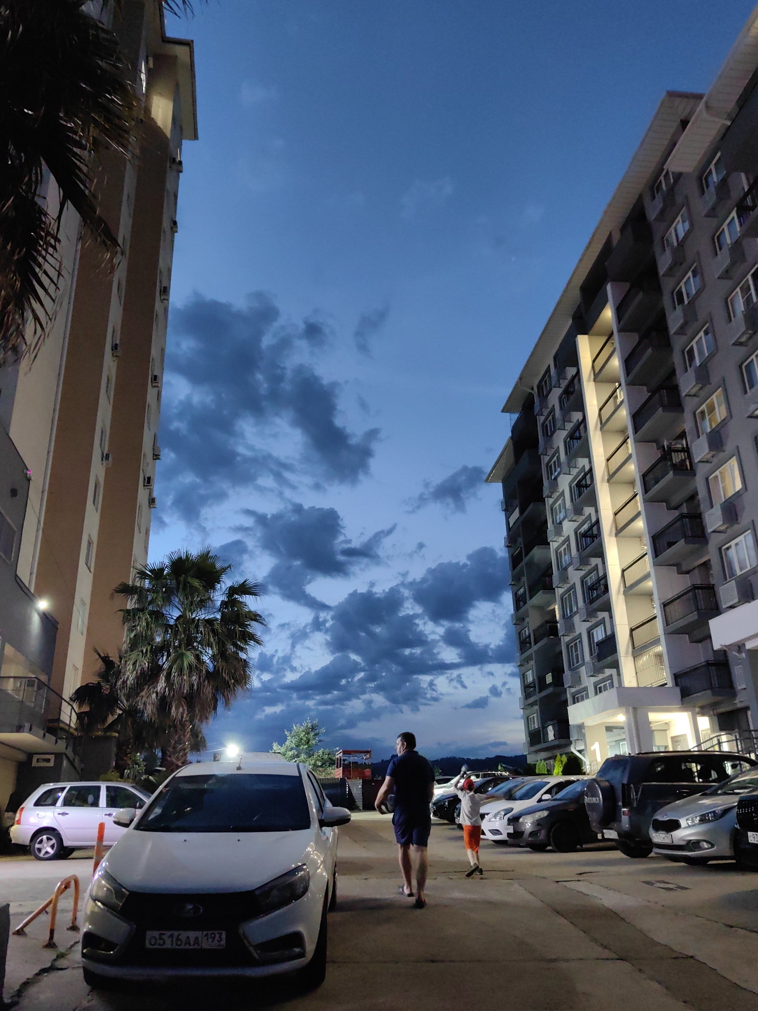 The image depicts a nighttime scene in a residential area, likely an apartment complex, in Sochi, Russia. Two modern, multi-story buildings are visible, one on the left and one on the right. The building on the left is lighter in color with visible balconies and air conditioning units. The building on the right is darker gray, also featuring numerous balconies and illuminated windows. Palm trees are present between the buildings and in the foreground, suggesting a subtropical climate.
Several cars are parked in a lot between the buildings. In the foreground, a white car with a Russian license plate is prominently displayed. Further back, a dark SUV and other cars are parked.
Two individuals are walking away from the camera towards the right side of the frame. The person in the foreground is an adult male wearing a dark blue t-shirt and navy blue shorts. The person behind him appears to be a child wearing an orange t-shirt and shorts. They are walking on a paved surface, which seems to be a parking lot or a pathway. The sky above is a deep blue, indicative of dusk or early night, with scattered clouds. The lighting suggests that artificial lights from the buildings and possibly streetlights are illuminating the scene.