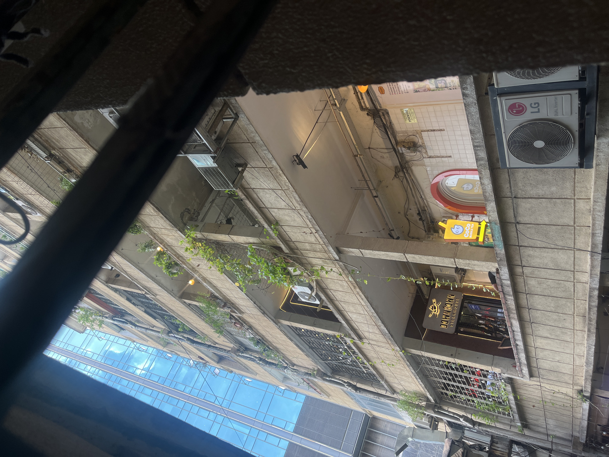 This is a view of an older apartment building in Ho Chi Minh City, Vietnam, seen from a lower vantage point, possibly through a window or balcony with a grill. The building is made of concrete with several balconies, some of which have plants growing on them. There are multiple air conditioning units visible on the exterior. One of the lower floors features the signage for "Downtown Steakhouse" and "Coco Bubble Tea." Adjacent to the apartment building is a modern building with a glass facade, reflecting the sky and clouds. The scene suggests an urban environment with a mix of old and new architecture.