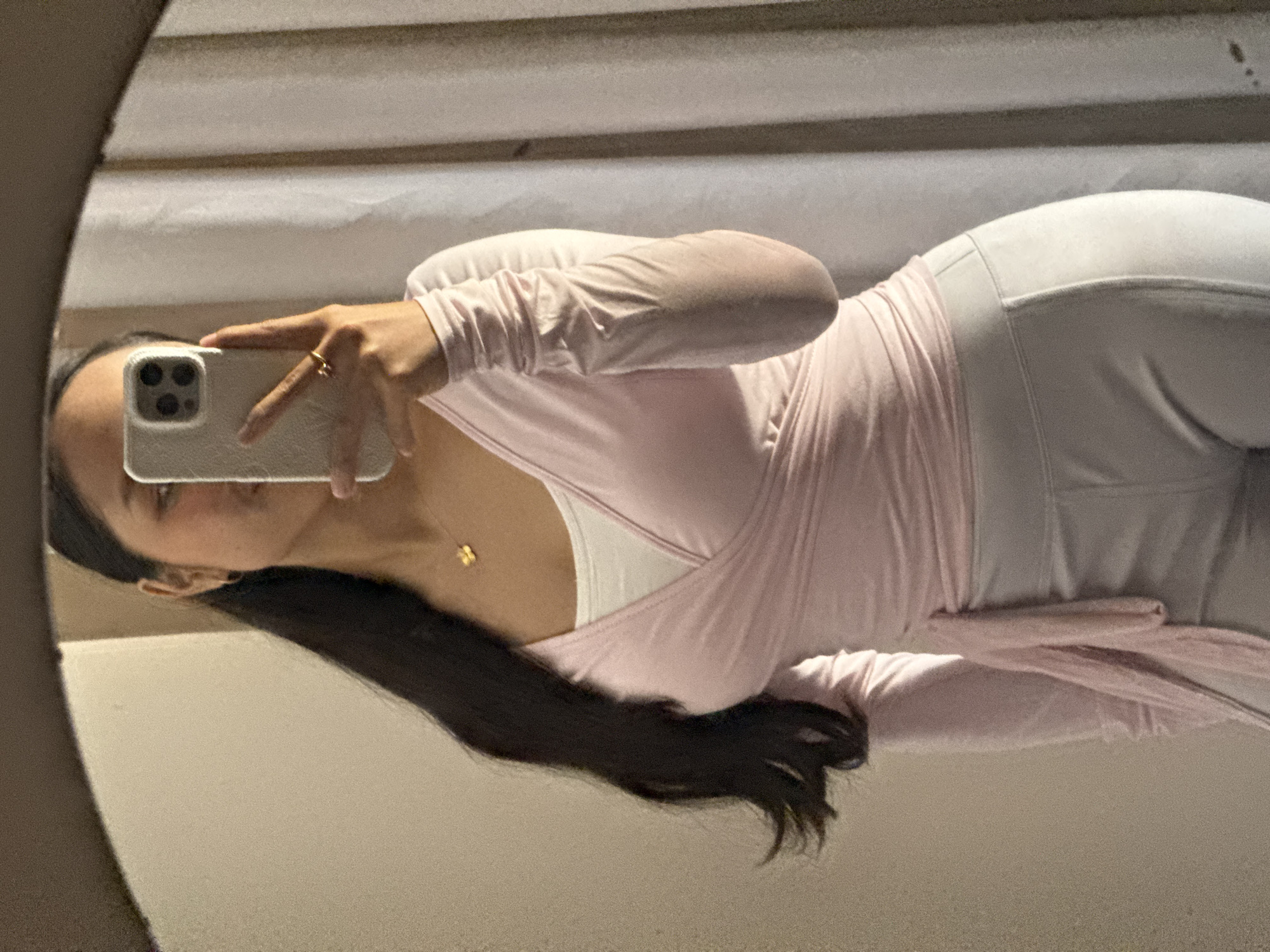 A woman is taking a selfie in a mirror. She is wearing a pink wrap top and white leggings. She has long dark hair and a gold necklace. She is holding a smartphone in her left hand and wearing a ring. The location is unknown, and there is no indication that it is in Davao City, Philippines. The background is a wall with horizontal panels.
