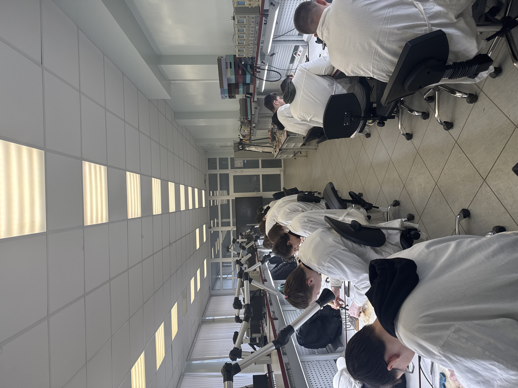A brightly lit, modern laboratory or workshop is depicted in Perm, Russia, filled with individuals engaged in technical work. Approximately ten young adults, dressed in white lab coats, are seated at individual workstations arranged in rows. They are focused on their tasks, leaning over work surfaces, suggesting activities like assembling, inspecting, or repairing electronic components.
Each workstation is equipped with an ergonomic, wheeled chair and a variety of specialized tools and electronic test equipment, including oscilloscopes and power supplies. Articulated arms, likely serving as fume extractors, magnifiers, or task lights, are prominent at each station, further emphasizing the detailed nature of the work being performed.
The room features a clean, functional design. The floor is covered with light-colored square tiles, and the ceiling is composed of grid panels embedded with numerous rectangular LED light fixtures, ensuring bright, uniform illumination throughout the space. The walls are also panelled in a light color, contributing to the sterile and professional atmosphere. At the far end, windows or glass doors are partially visible, reflecting light. The scene conveys a sense of diligent study or professional activity in a well-organized, technical environment. There is no visible text clearly discernible in the image.