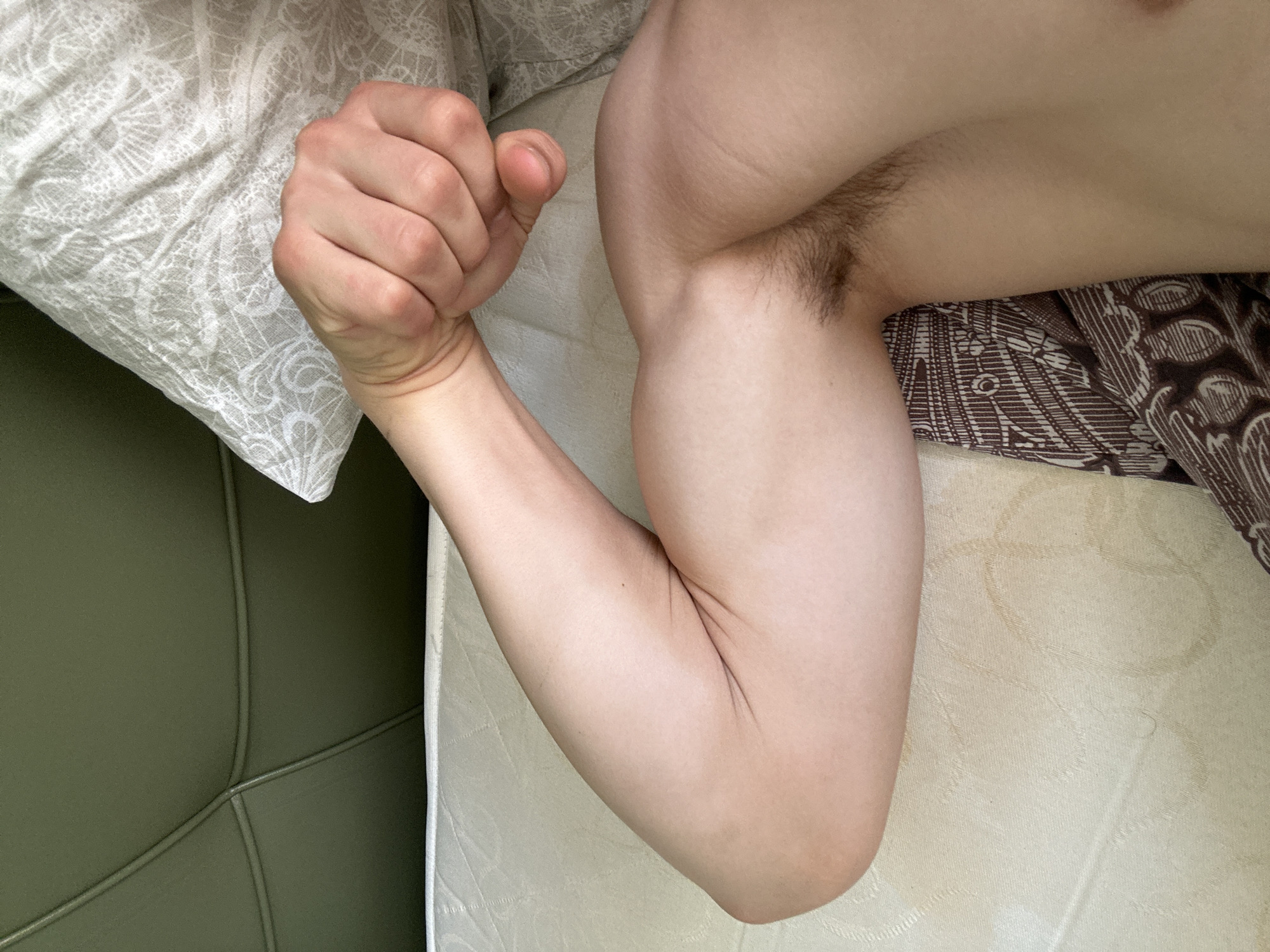 An upper arm and clenched fist are visible in the image. The arm is positioned on a surface with patterned bedding. The arm has some underarm hair. A pillow is situated near the clenched fist.