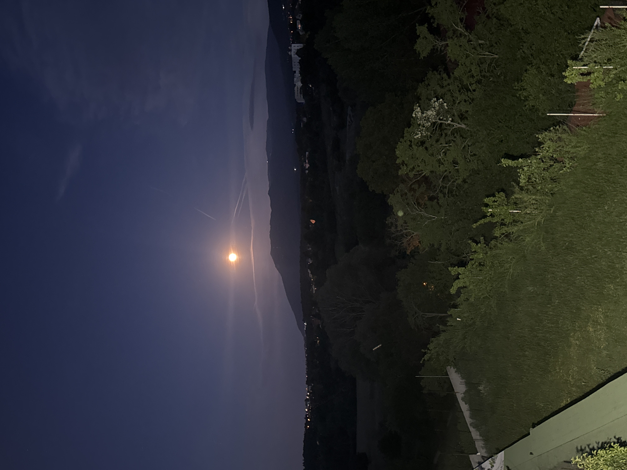 The image depicts a night scene in an unknown city. A bright moon dominates the dark blue sky, with contrails of an aircraft visible nearby. Below the horizon lies a mountain range, illuminated by scattered lights suggesting a populated area along its base. The foreground is filled with trees and foliage, and a partial view of a green roof is visible at the bottom right corner of the image. White lines can be seen on the ground in the foreground.