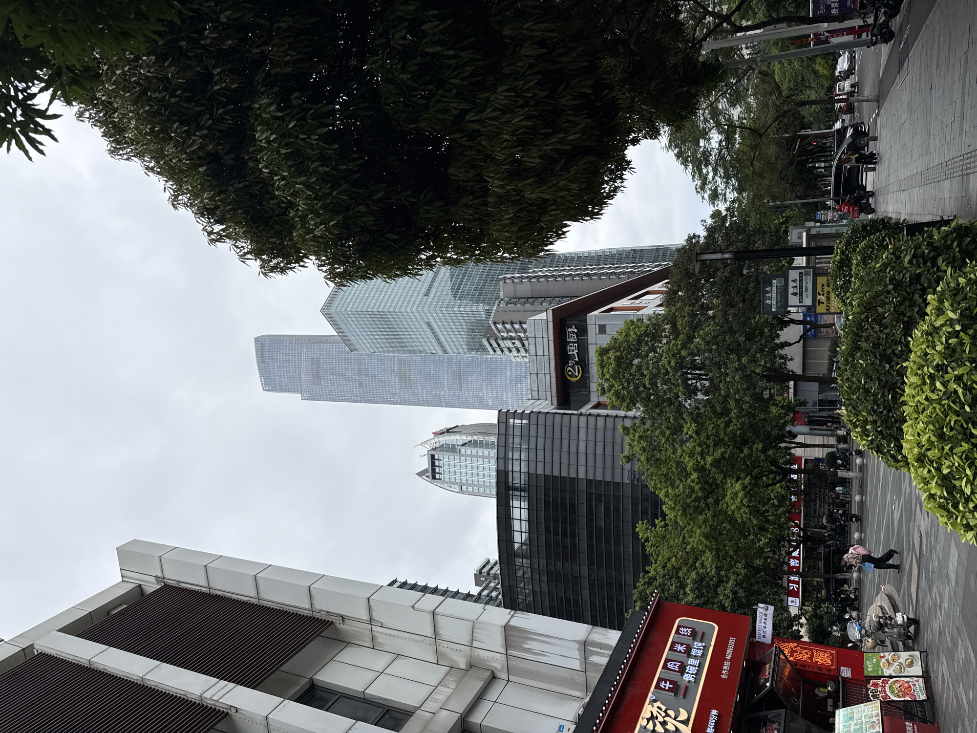 The image depicts a street scene in Tianhe District, China, under a cloudy sky. In the foreground, a red storefront with Chinese characters and illuminated signs is visible on the left. Adjacent to it, a person is walking with a backpack. Further down the sidewalk, several trees line the street, interspersed with modern buildings. The middle ground features a row of trees and the facade of a building with red accents and the text "2号厨房" (Number 2 Kitchen). Beyond this, several tall skyscrapers dominate the background, with a very tall, slender skyscraper in the center. The street is paved with gray tiles, and a few vehicles, including motorcycles and cars, are present on the road. Lush green bushes are planted along the edge of the sidewalk.