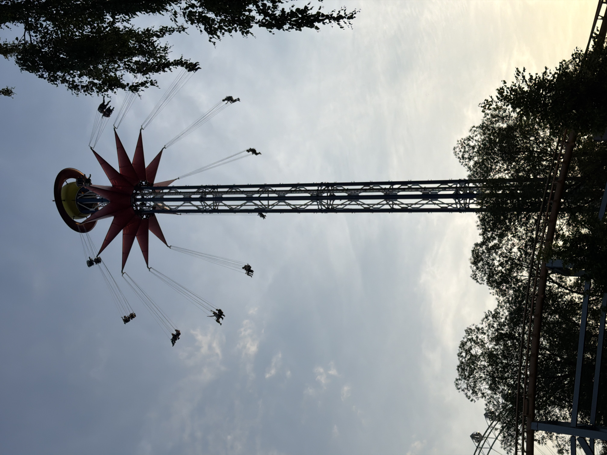 This is a low-angle, vertical shot of a swing ride at an amusement park, likely located in Saint Petersburg, Russia. The sky is overcast with patches of brighter clouds, suggesting it's either early morning or late afternoon. The swing ride itself is a prominent red, star-shaped structure at the top, with a yellow ring around its central hub. Long metal cables extend outwards and downwards, from which numerous individual swings are suspended. Several people are visible on the swings, appearing as dark silhouettes against the sky. They are in motion, suggesting the ride is in operation. The overall impression is one of a fun amusement park experience on a somewhat subdued day. Trees and parts of other amusement park structures are visible in the background, adding context to the setting.