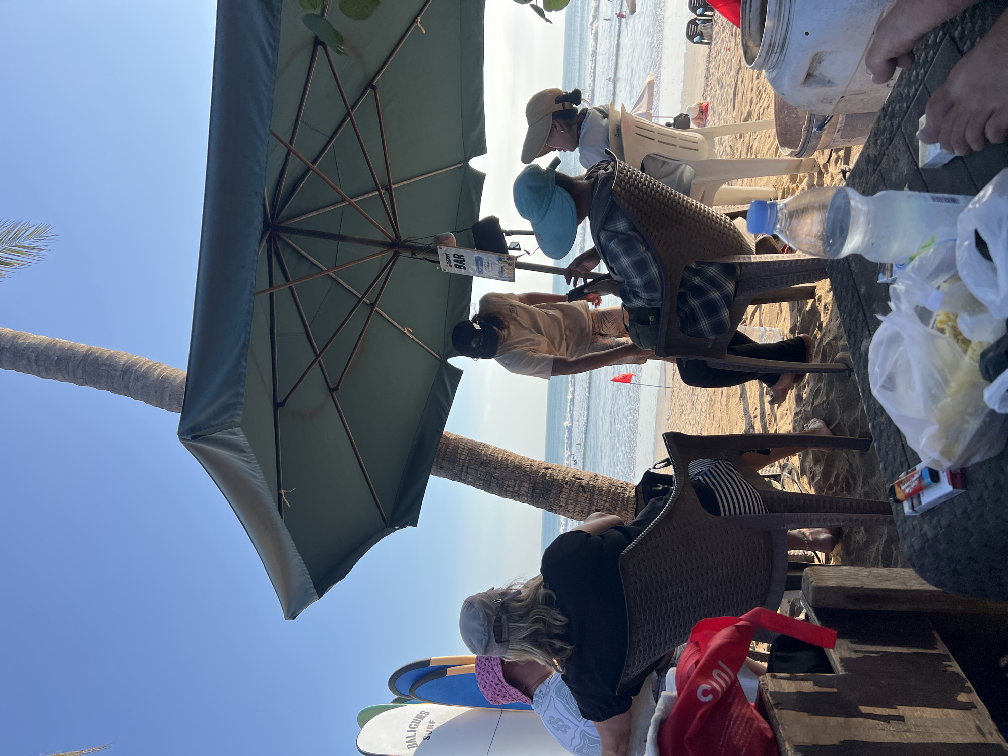 In Kuta, Indonesia, on a sandy beach, a group of people are gathered under a large green umbrella, enjoying a sunny day. Several surfboards are propped up behind them, suggesting proximity to the ocean. The people are seated on chairs, some facing the water, while others are engaged in conversation or looking at their phones. A sign for "BAR CHERRY" hangs from the umbrella's pole. A bottle of water and some snacks are visible on a table in the foreground. The ocean waves are gentle, and a few people can be seen swimming in the distance. A palm tree trunk is also visible, adding to the tropical beach ambiance.