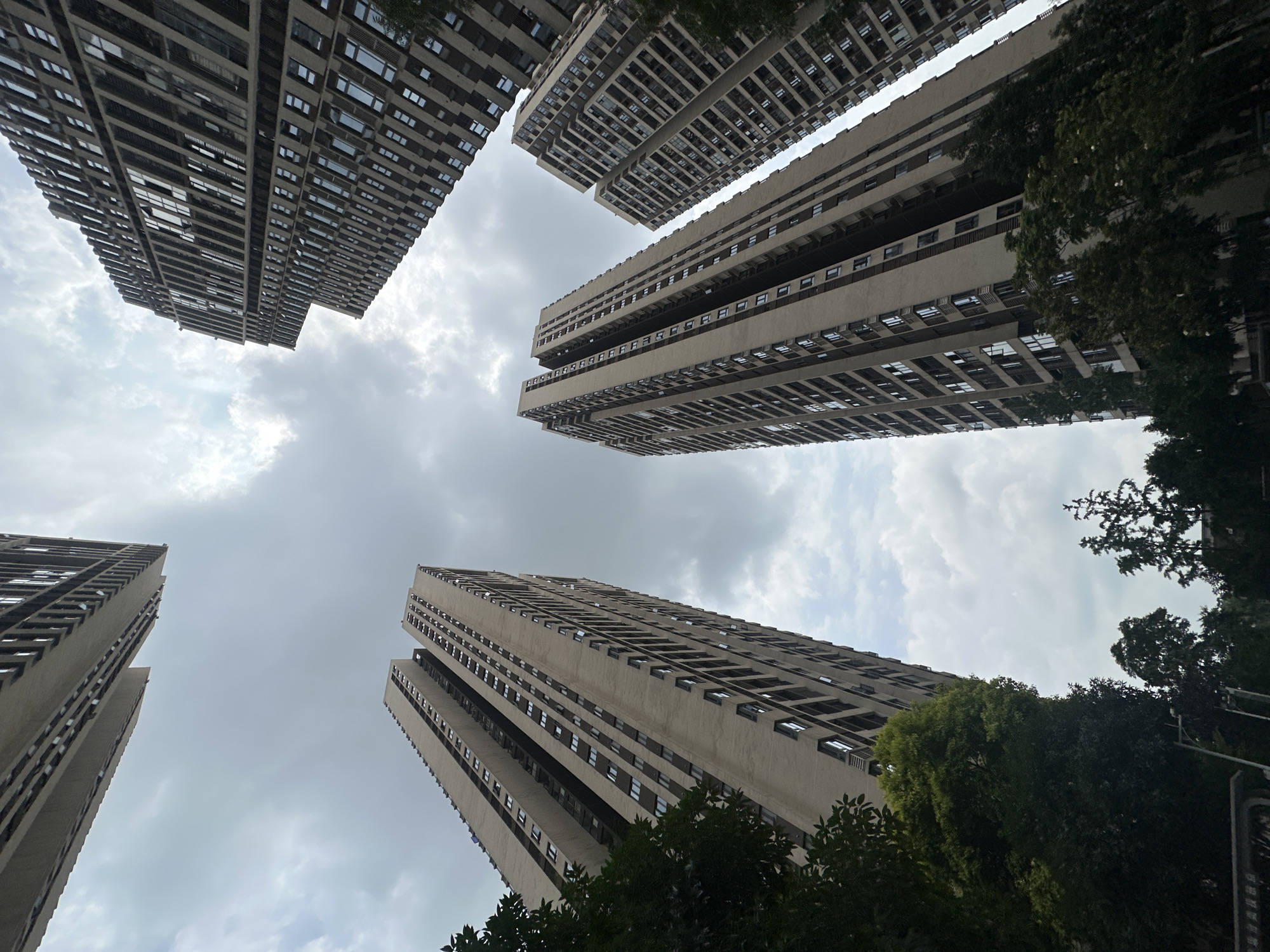 The image, taken from a low angle, shows multiple high-rise apartment buildings against a cloudy sky. The buildings are mostly beige or light gray with rows of windows and horizontal architectural elements. Some buildings are partly obscured by trees with green foliage. The location is in the city of Chongqing High-tech Zone, China. No people are visible.