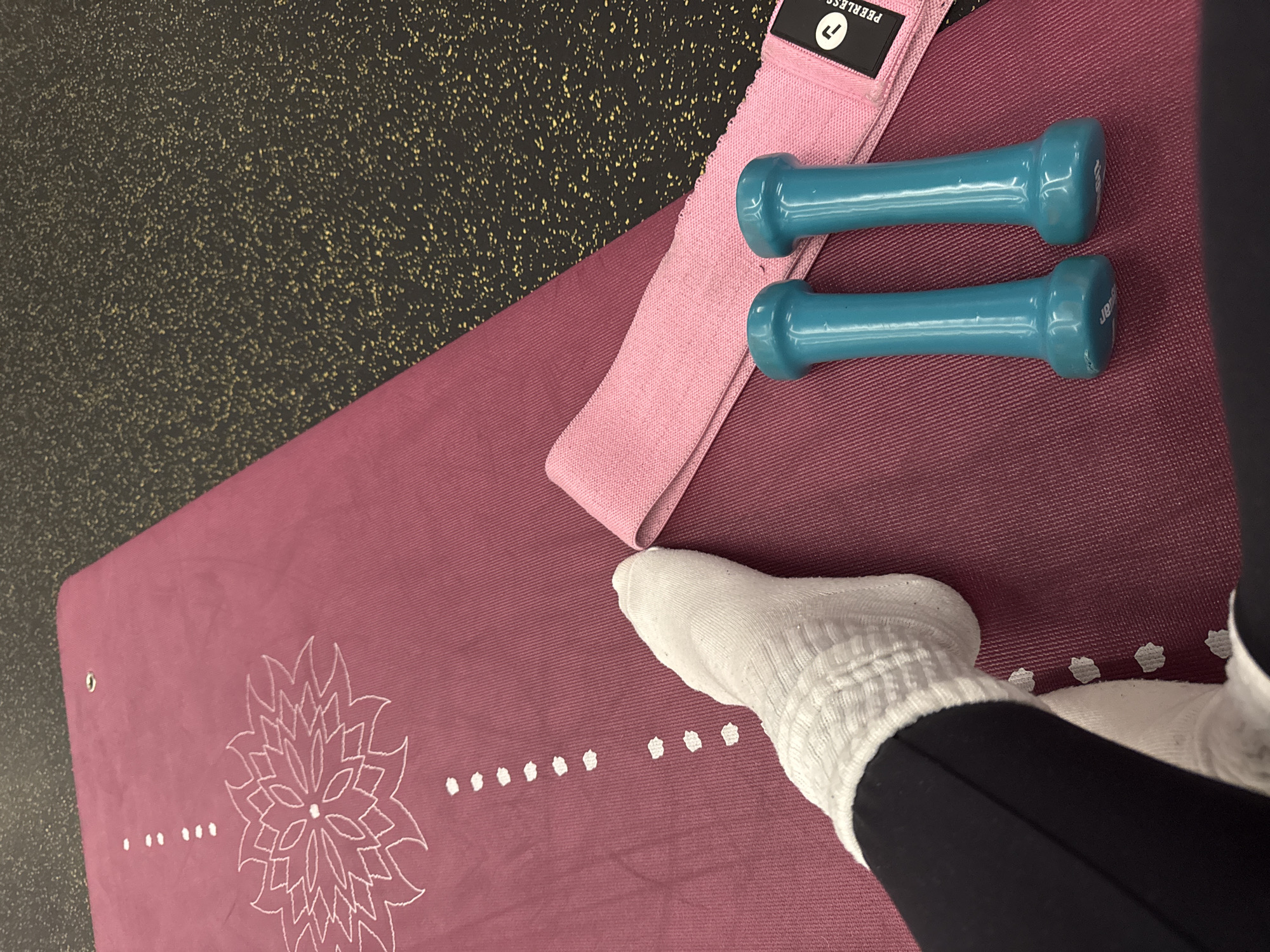 The image depicts a section of a maroon yoga mat with a white floral design and a row of white dots. A pink resistance band and a pair of teal dumbbells are on the mat. A person's foot and lower leg are visible, with the foot wearing a white sock and the leg covered by black leggings. The setting appears to be indoors, possibly a gym or home workout space. The floor visible around the mat is black with yellow flecks. There is no evidence to suggest the location is Sofia, Bulgaria.