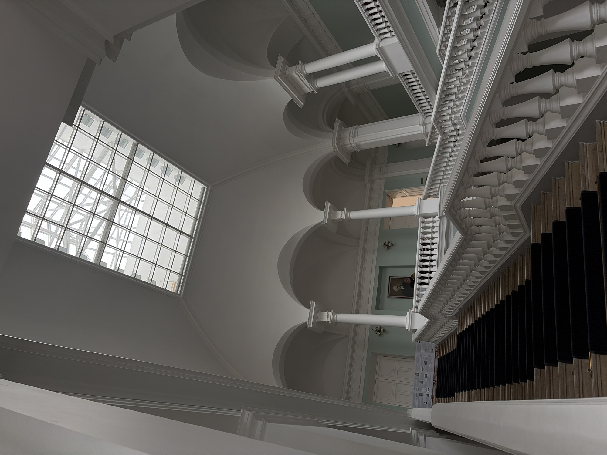 This is an interior photograph of the Grand Staircase in the Russian Museum in Saint Petersburg, Russia. The image captures the expansive scale of the staircase with its white balustrades and a black carpet runner. Above the staircase, a large glass skylight illuminates the space. The architecture features classical elements, including white columns, arched ceilings, and decorative molding. On the upper landing, a portrait of a man in a suit hangs on a pale blue wall. Two individuals are visible on the upper landing, one standing near the portrait and another leaning on the balustrade. The scene conveys a sense of grandeur and historical significance, typical of the museum's architecture.