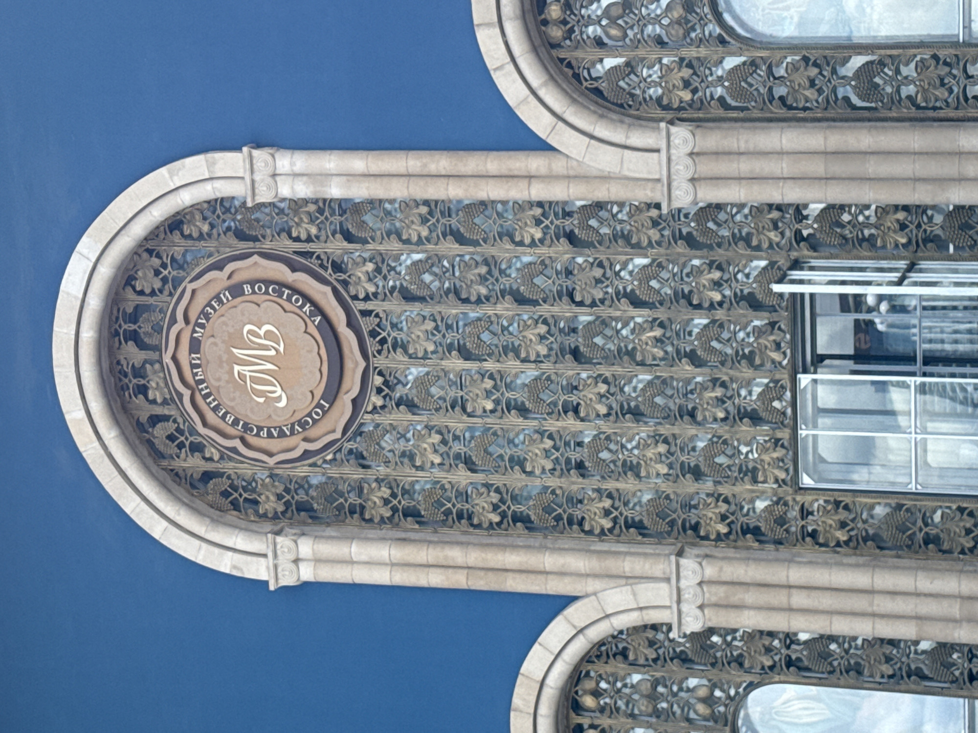 The image shows an architectural detail of a building with a blue sky background. The building features ornate stonework, including arched windows with intricate metal grilles. The grilles have a repeating pattern of stylized leaves and grape clusters. Above the grilles, there are decorative stone moldings.
A prominent circular emblem is visible on the left side of the building's facade. This emblem contains Cyrillic text and a stylized monogram. The text, when deciphered from the circular inscription, reads "ГОСУДАРСТВЕННЫЙ МУЗЕЙ ВОСТОКА" (State Museum of the East). The monogram inside the emblem appears to be "MB".
There are windows visible in the background, suggesting this is an exterior shot of a building. The light in the image appears to be diffused, hinting at an overcast day or late afternoon. The overall impression is of a building with significant historical or cultural importance, given the elaborate ornamentation and the presence of a museum's insignia. The architecture suggests a style that might be found in Moscow, Russia, as indicated by the location context. No people are clearly visible in the image.