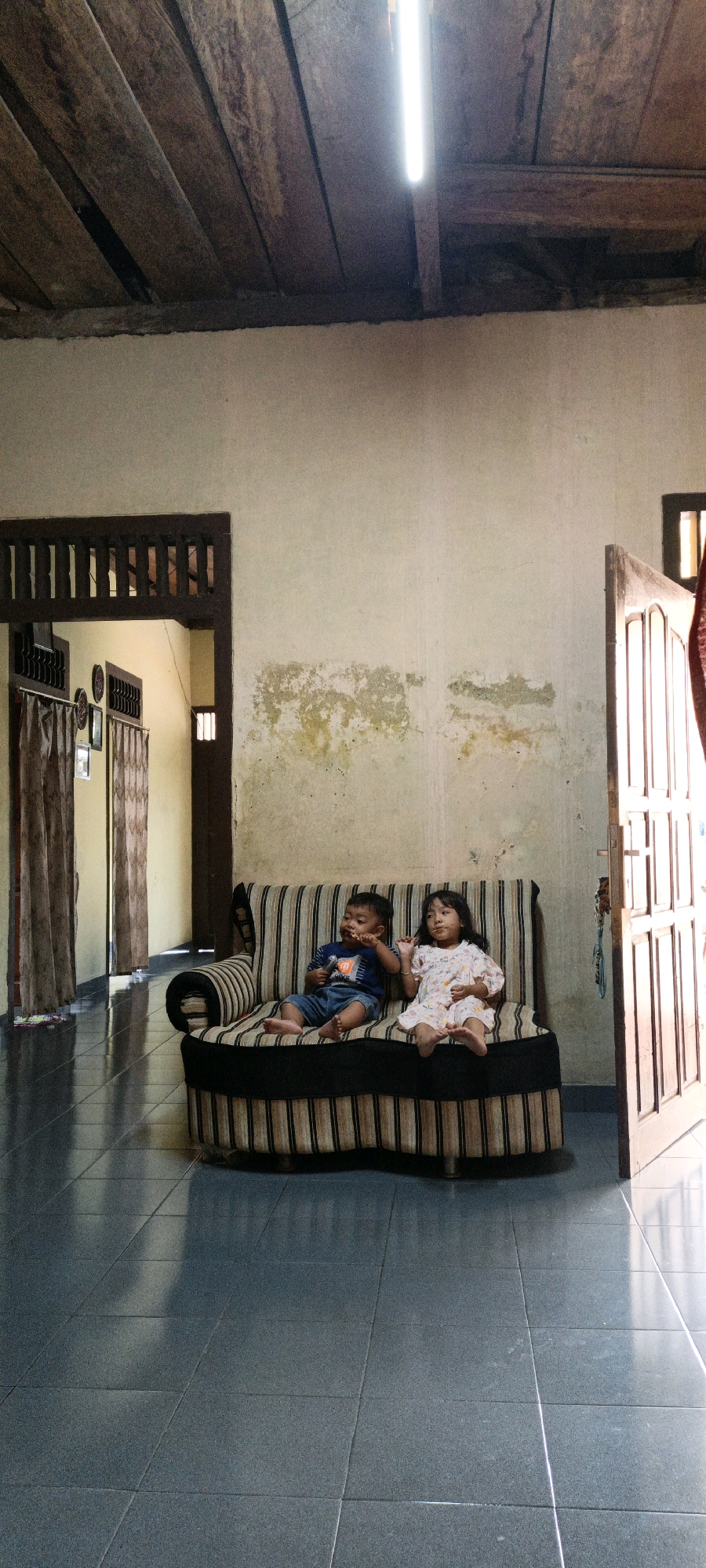 In a dwelling in Bunut, Indonesia, two young children, a boy and a girl, are seated on a striped sofa. The boy, wearing a blue t-shirt and denim shorts, is holding food to his mouth. The girl, dressed in a floral patterned dress, looks towards the viewer with food on her fingers. They are positioned in a room with tiled flooring and walls that exhibit signs of peeling paint. The room is illuminated by a fluorescent light fixture attached to a wooden ceiling. Open doorways reveal glimpses of adjoining areas, including a hallway with curtains and another doorway. The overall atmosphere suggests a simple, domestic setting.