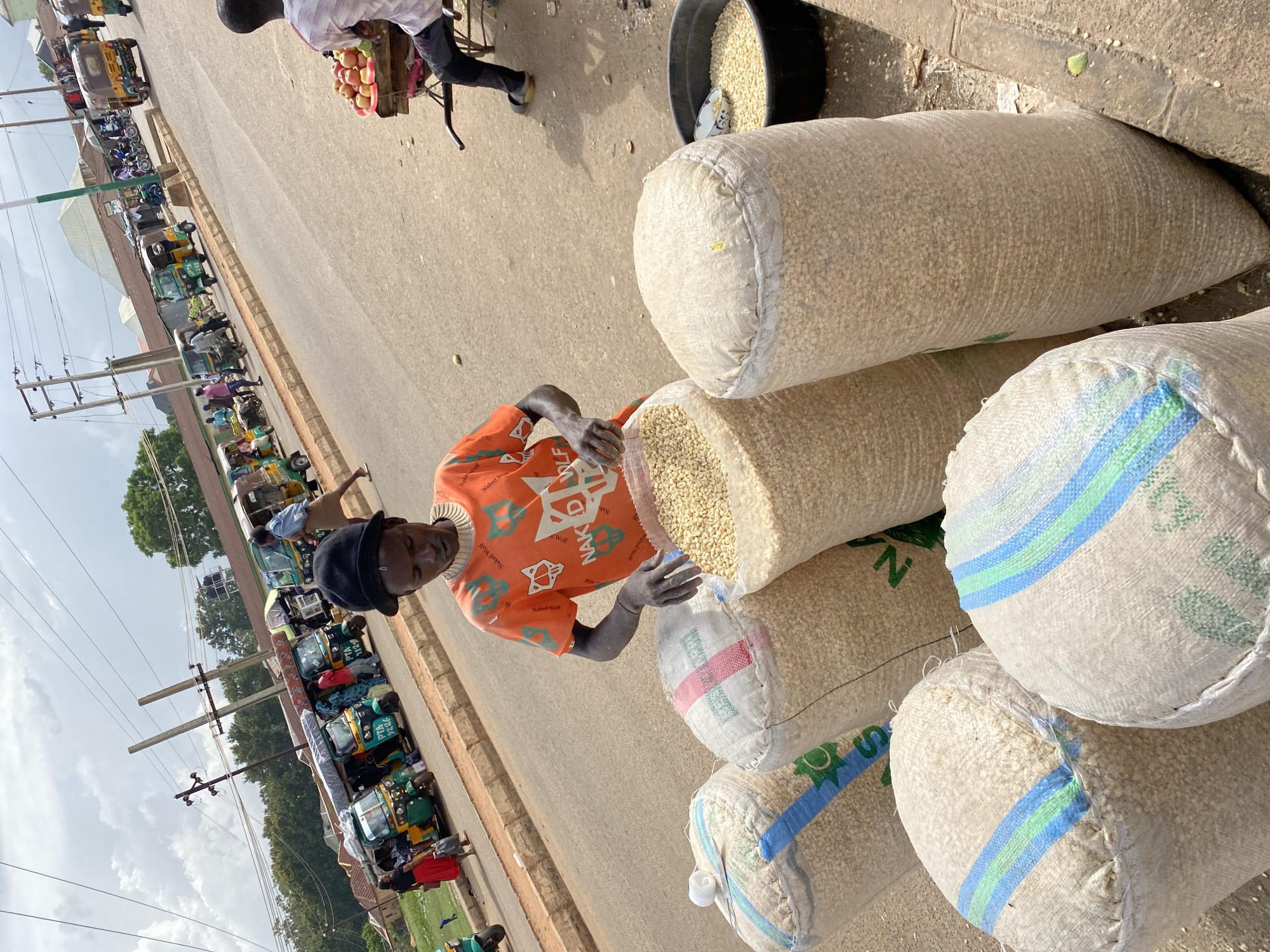 In Bukuru, Nigeria, a young male vendor, wearing an orange t-shirt with a "Naked Wolf" graphic and a black cap, is positioned at the roadside. He is interacting with large sacks filled with what appears to be dried corn kernels. Three sacks are prominently visible in the foreground, one of which the vendor is tending to. A black basin containing more corn kernels is situated to the right. The background shows a busy street with several three-wheeled auto-rickshaws (Keke Napep) and a motorcycle, indicating a bustling market or transport hub. The sky is partly cloudy, and the overall environment suggests an outdoor market scene in a Nigerian city.
